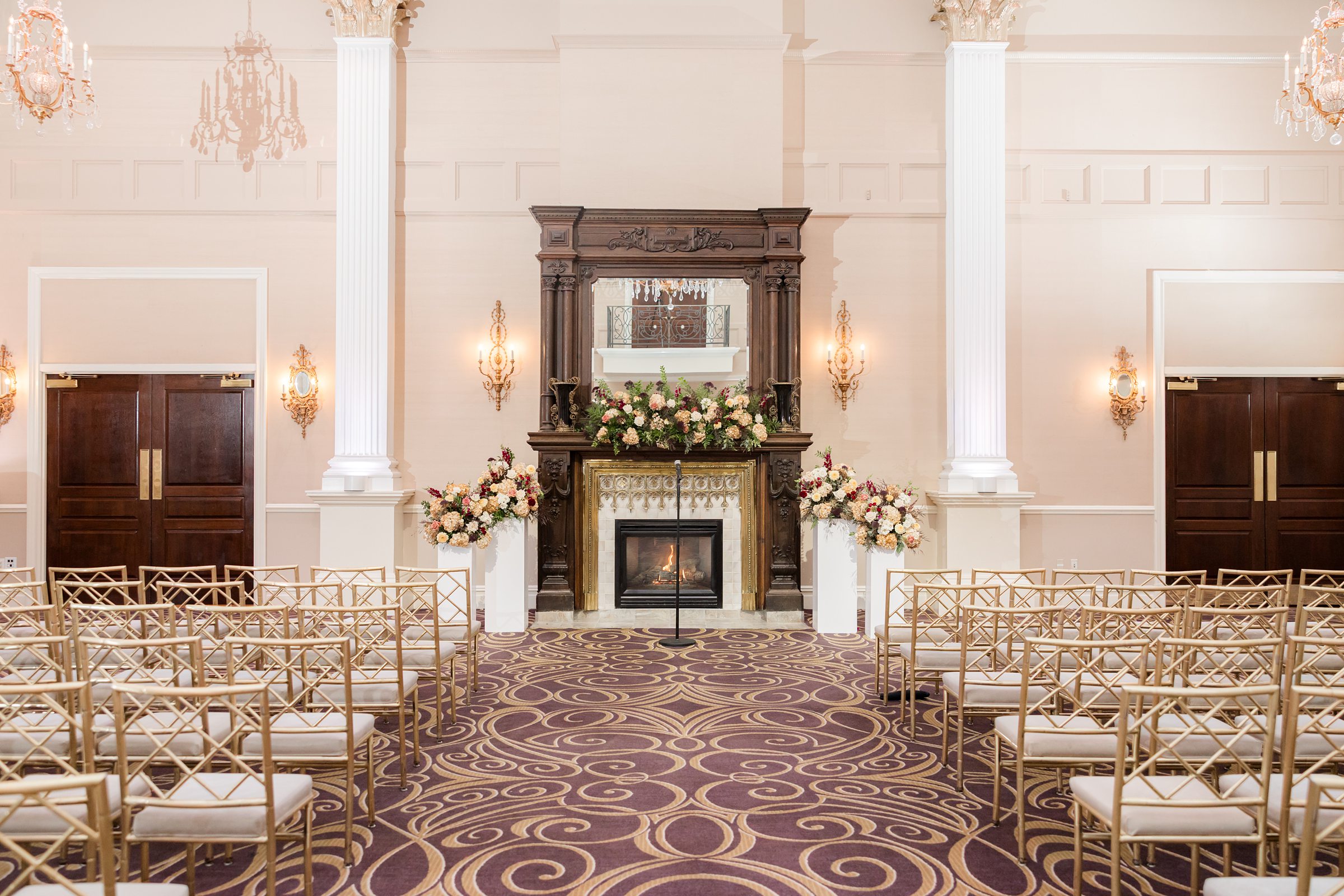 Elegant wedding ceremony space with gold chairs, a grand fireplace, and soft floral arrangements under warm chandelier light.