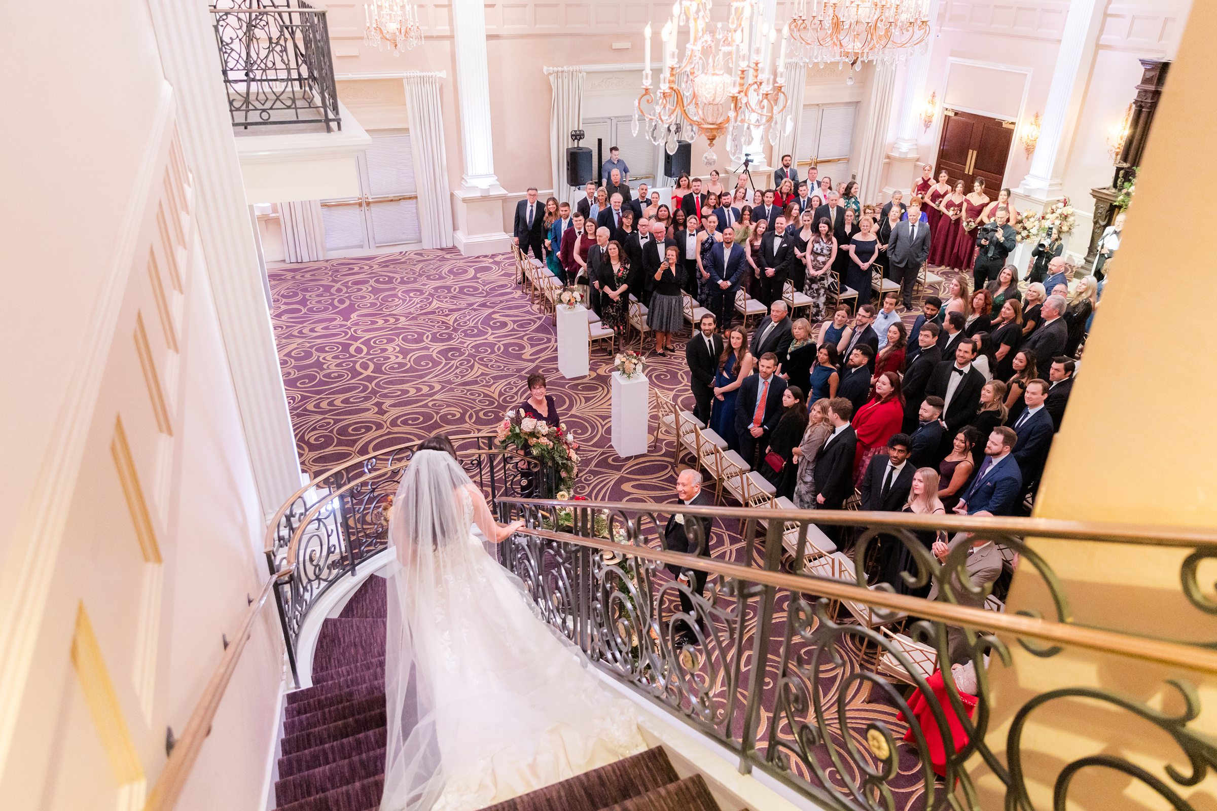 Bride descends a grand staircase toward her ceremony as guests look on in an elegant ballroom.