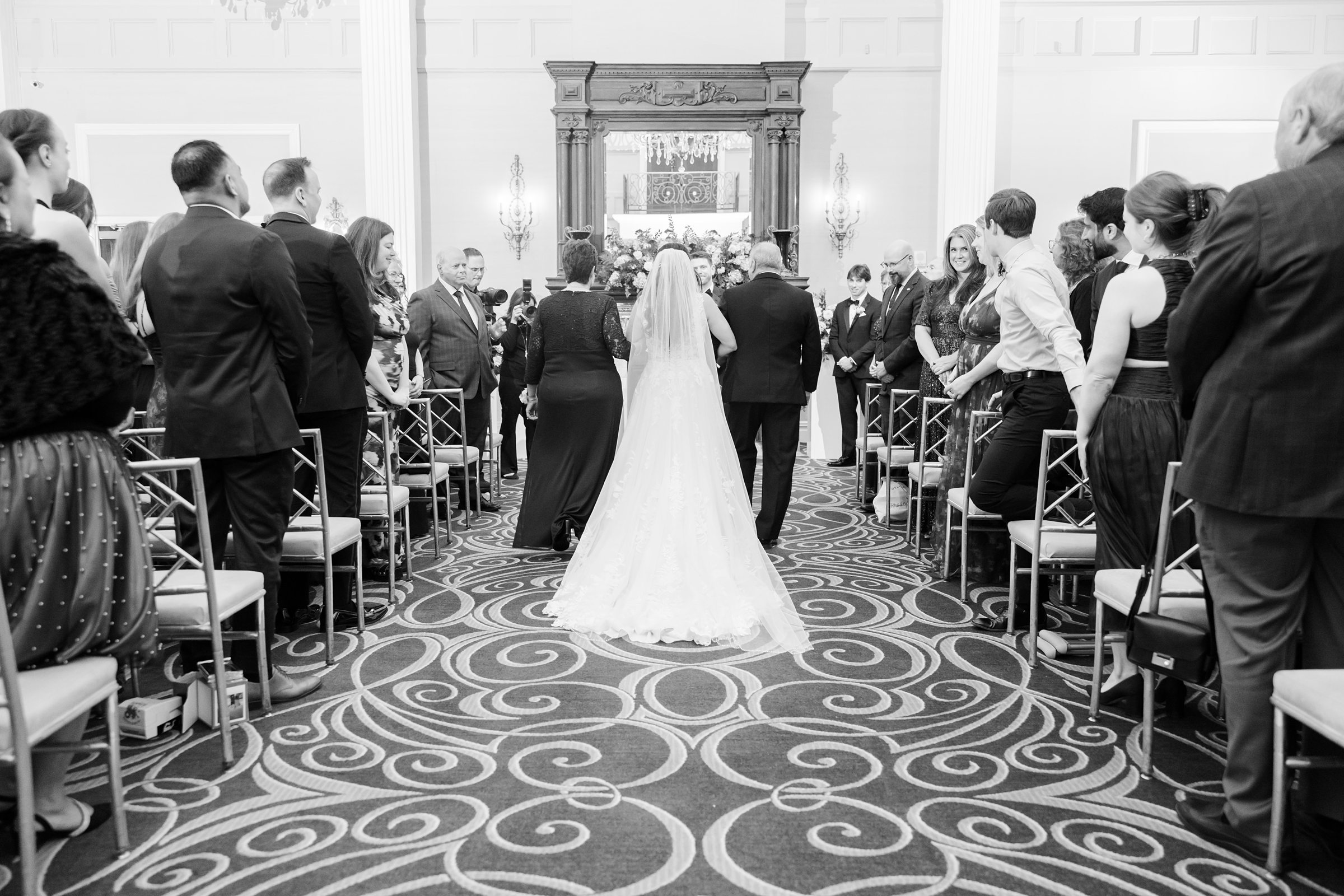 Bride walks down the aisle with her parents, a tender and emotional moment as loved ones look on with warmth and admiration