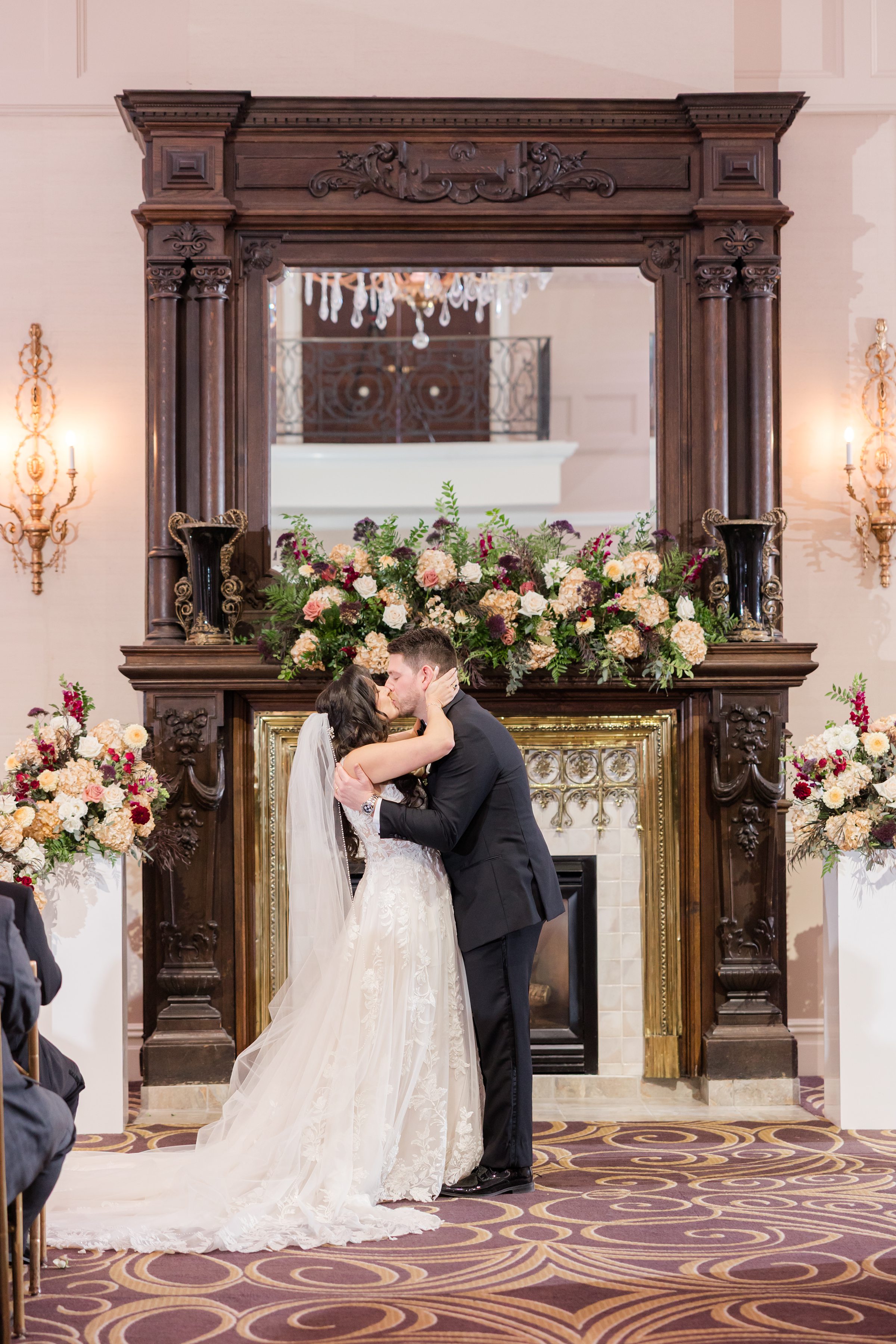 Bride and groom share a romantic kiss in front of a grand fireplace adorned with lush florals, celebrating their just married moment.