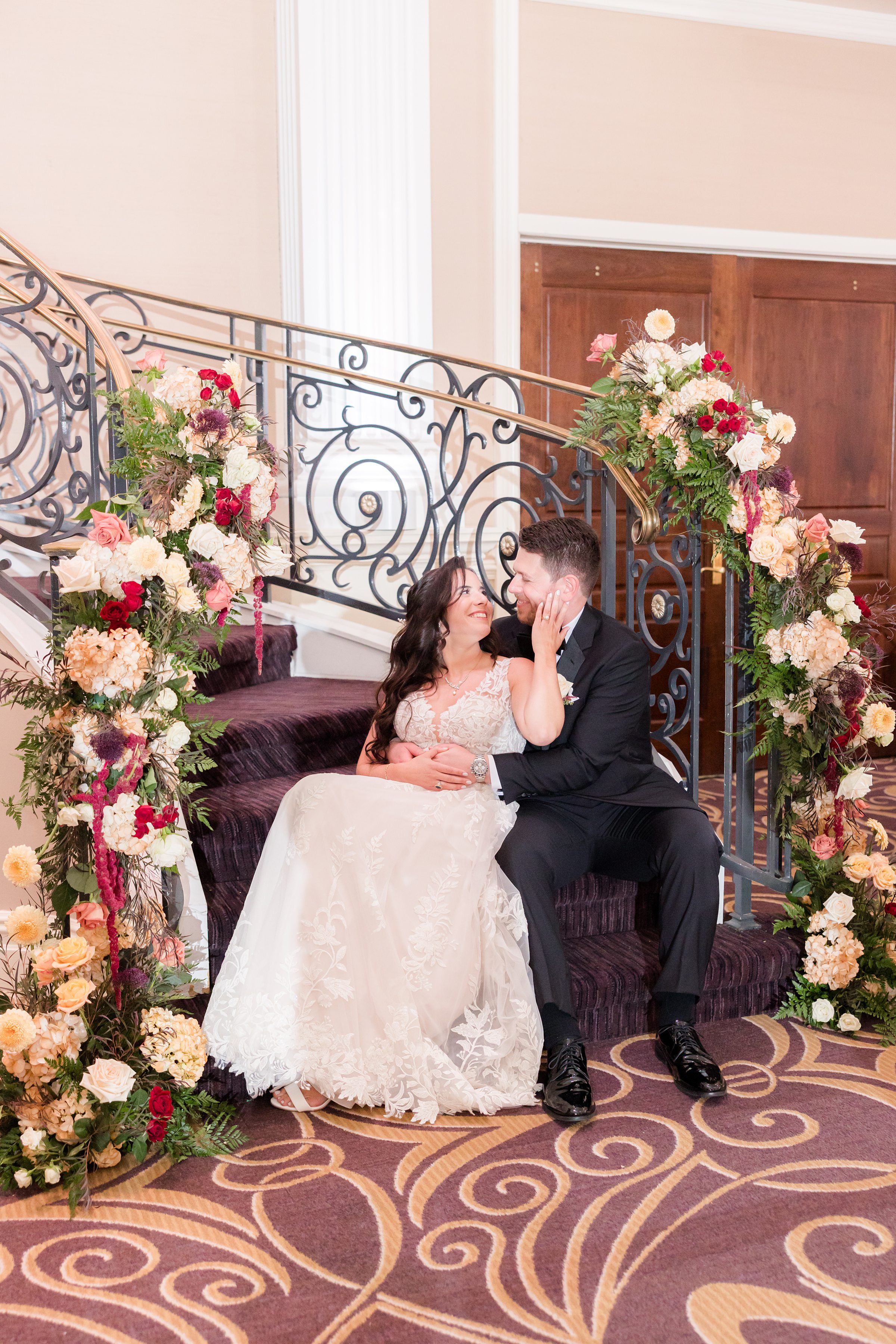 Bride and groom sit close on a staircase surrounded by flowers, sharing a tender, intimate moment and smiling at each other