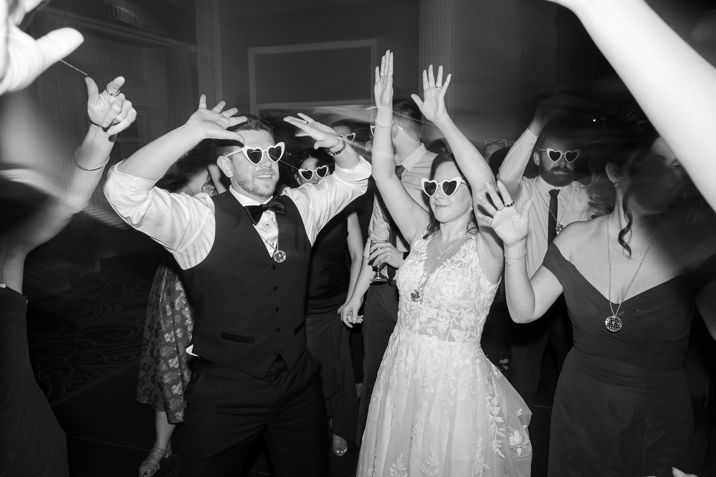 Bride and groom dance together on a crowded dance floor, raising their hands in celebration as guests join in around them