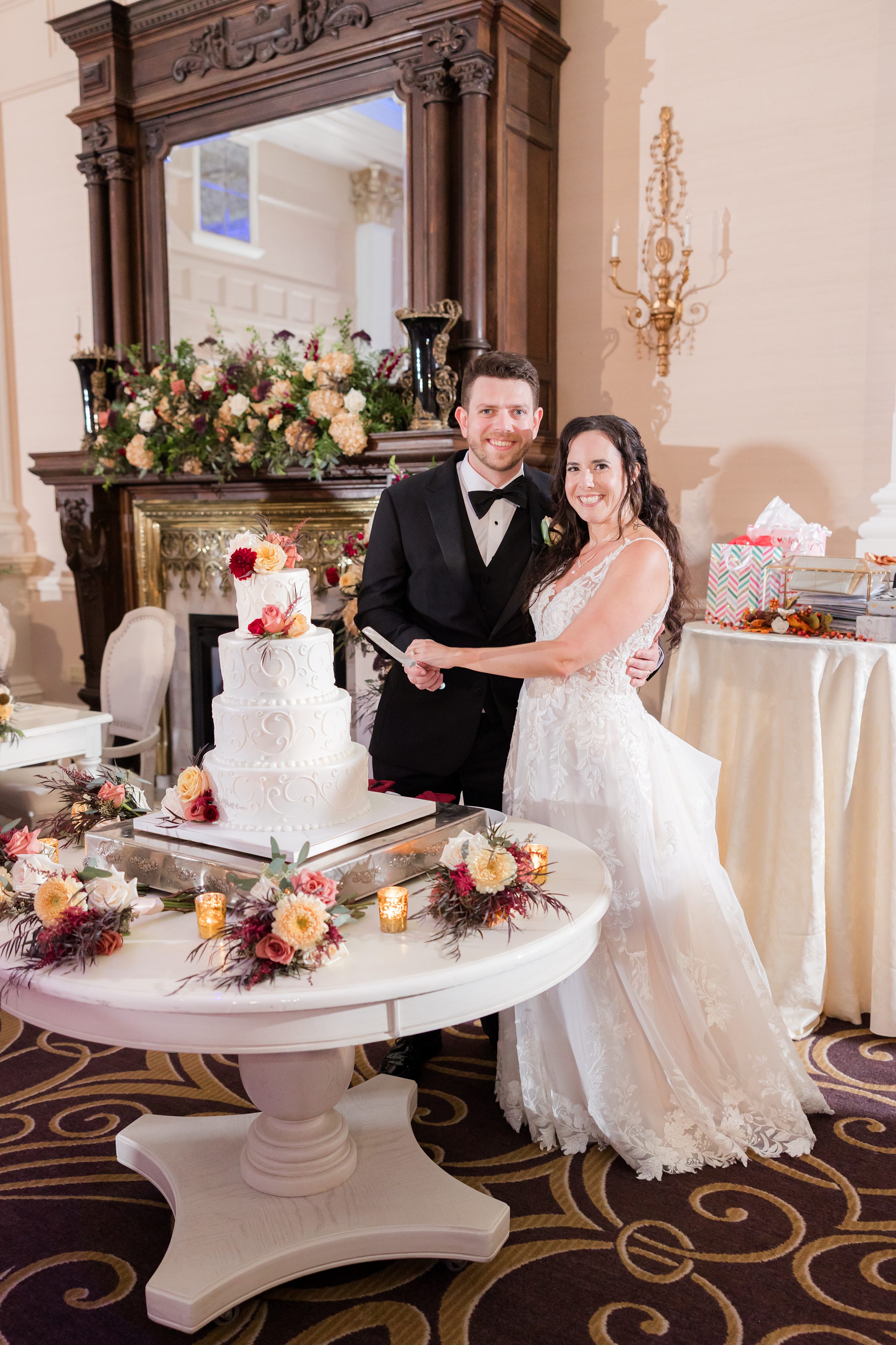 Bride and groom stand together beside their wedding cake, smiling warmly as they hold the cake knife during their reception