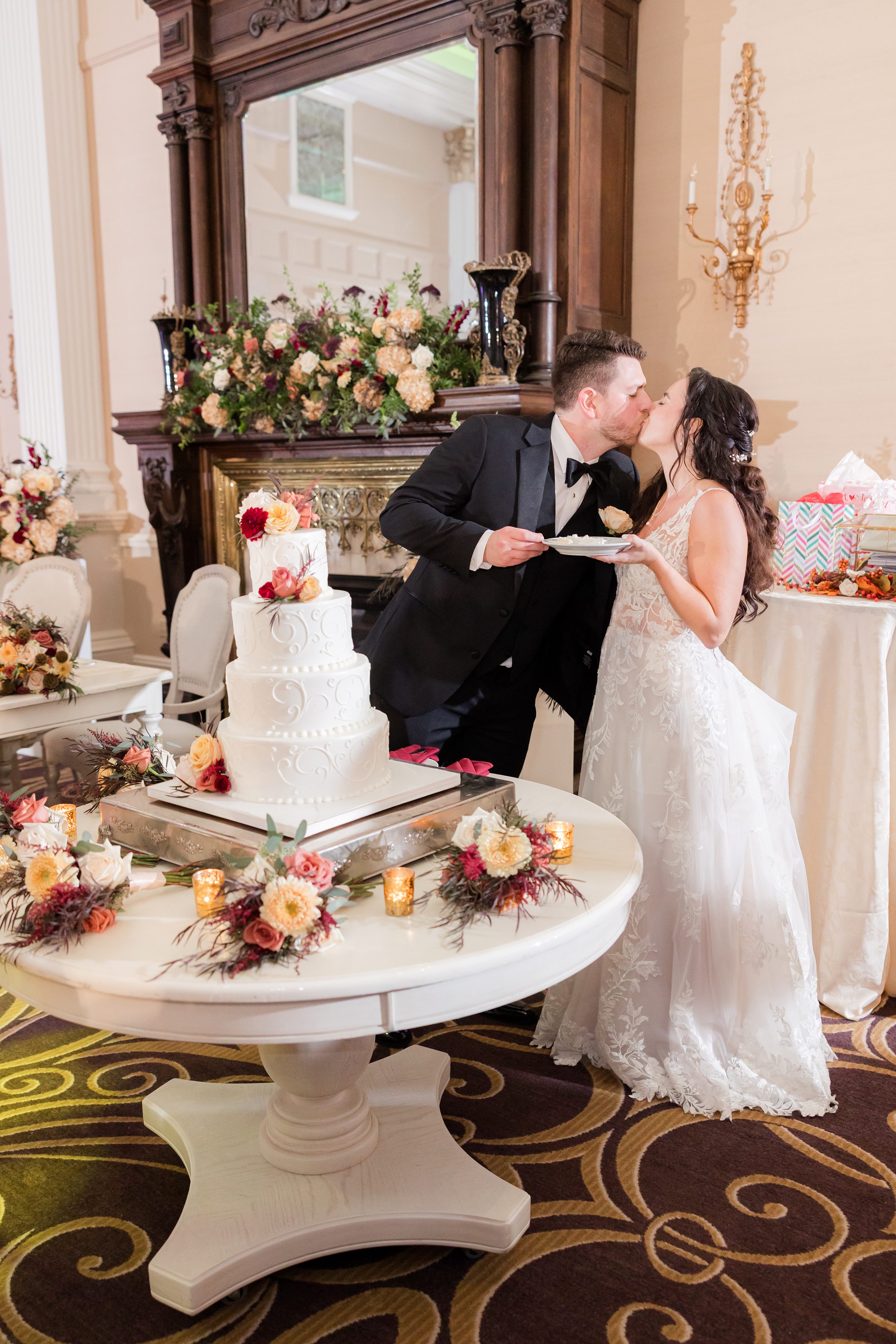 Newlyweds share a sweet kiss while feeding each other cake beside a floral adorned wedding cake, surrounded by warm candlelight and elegant décor. Fireplace night portrait