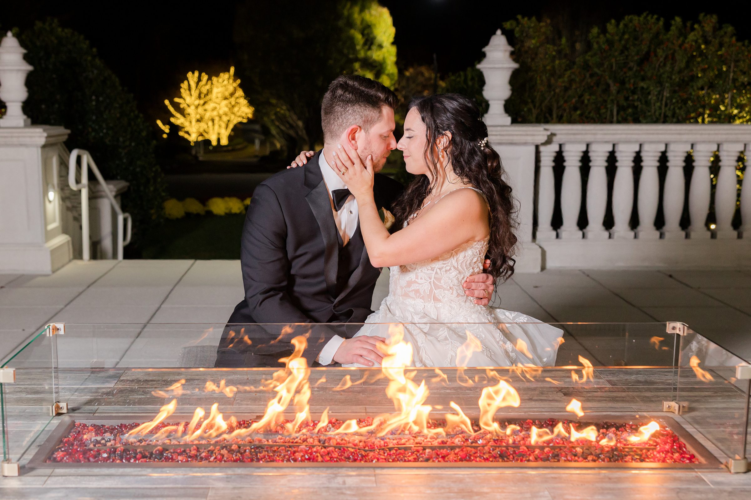 Bride and groom sits close by a glowing outdoor fire, gazing into each other’s eyes with quiet intimacy under the night sky
