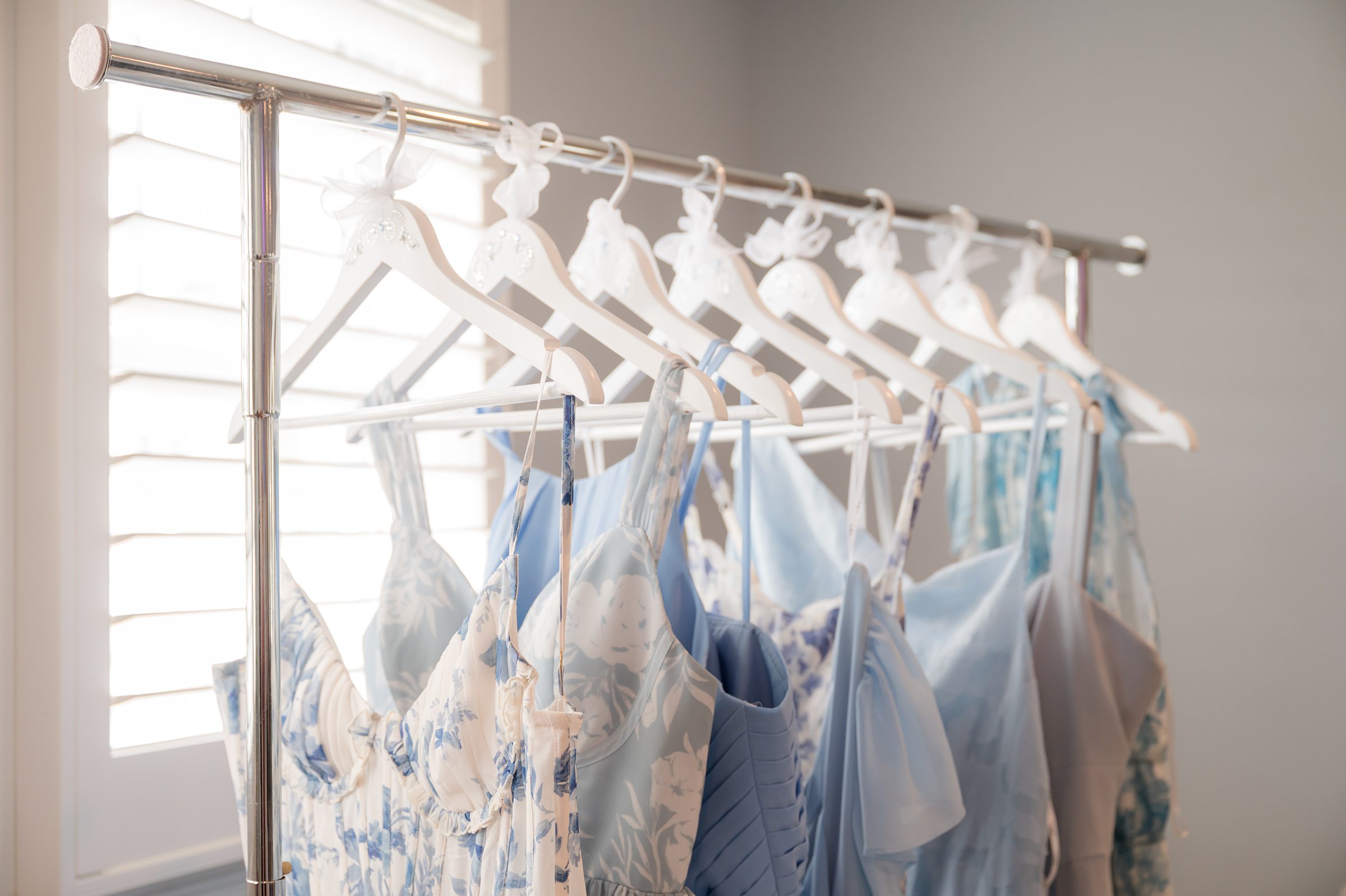 Soft blue bridesmaid dresses hanging neatly on a rack by a sunlit window, creating a calm and romantic getting ready scene.