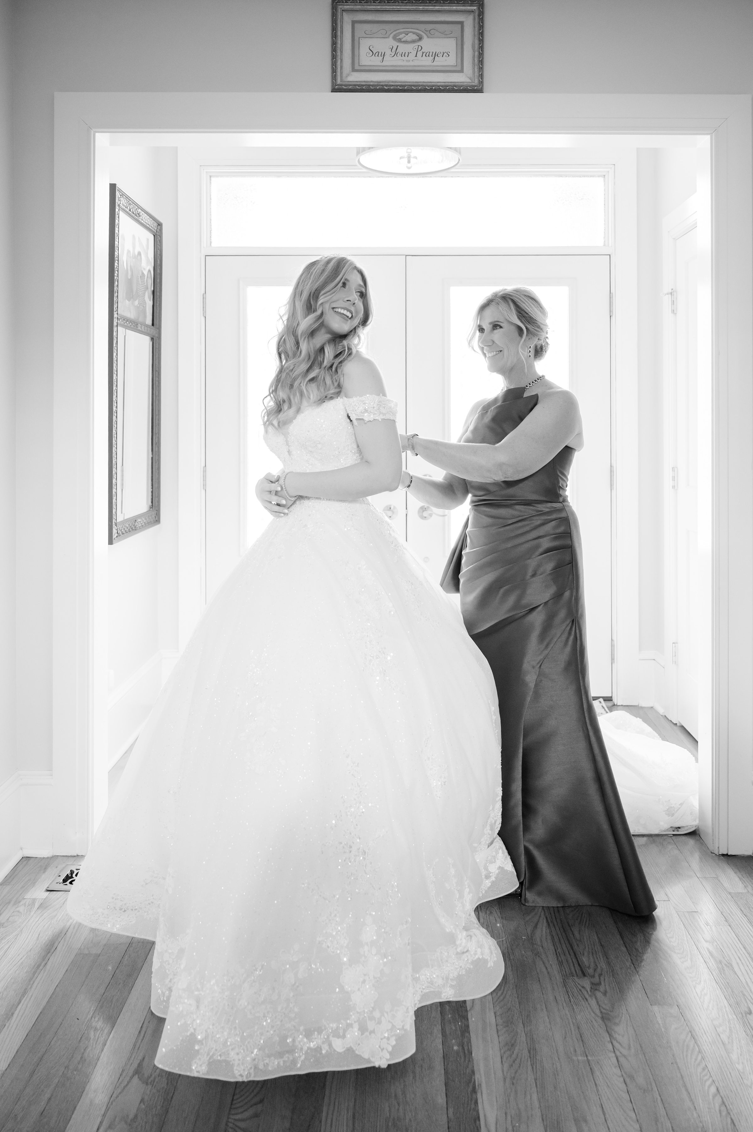 Bride and her mother sharing a sweet moment as they adjust her wedding dress in soft, glowing light.