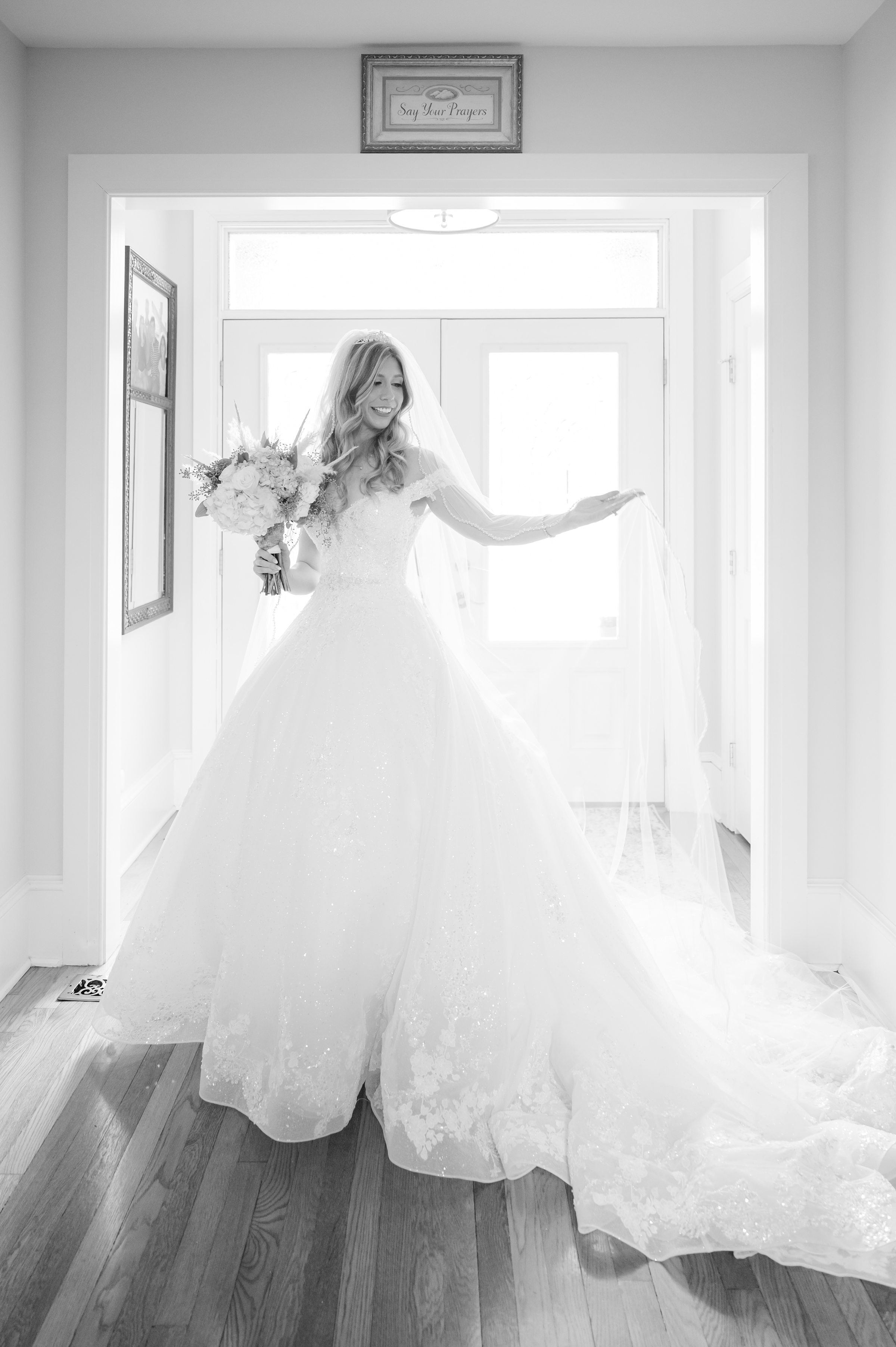 Bride holding her bouquet and veil, softly lit as she stands in a doorway, savoring a quiet, joyful moment before the ceremony.