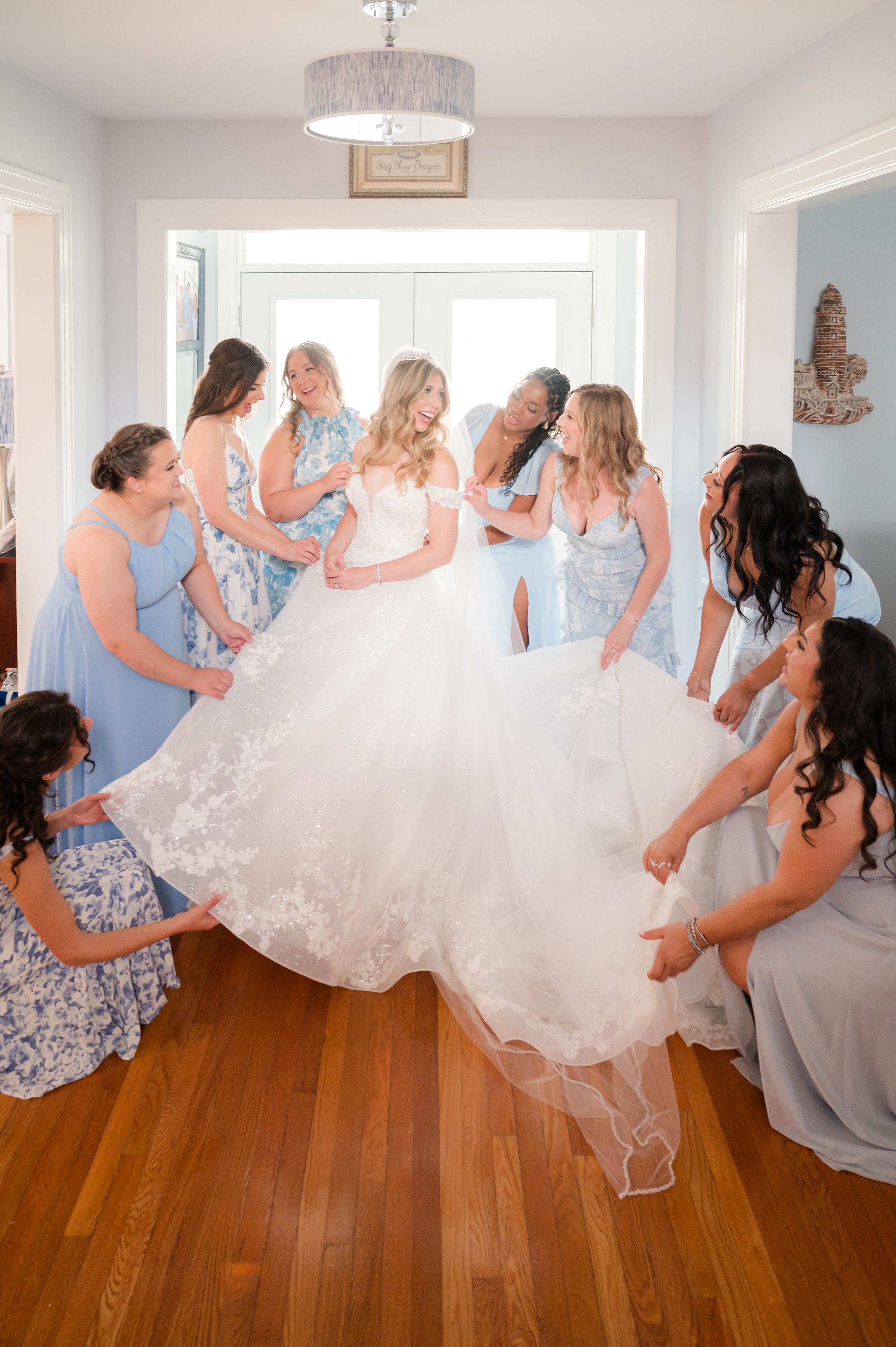 Joyful bride surrounded by her bridesmaids in soft blue dresses, sharing laughter as they help adjust her gown in a bright, love-filled room.