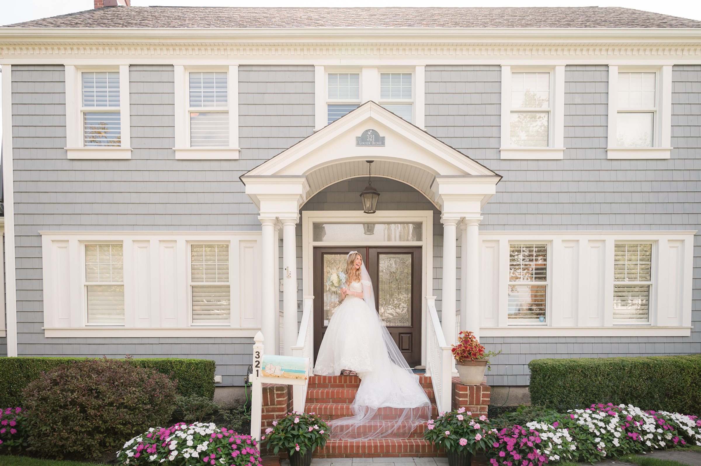 Bride standing on the front steps of a charming seaside home, her gown flowing as she pauses in a peaceful, romantic moment before the celebration begins.