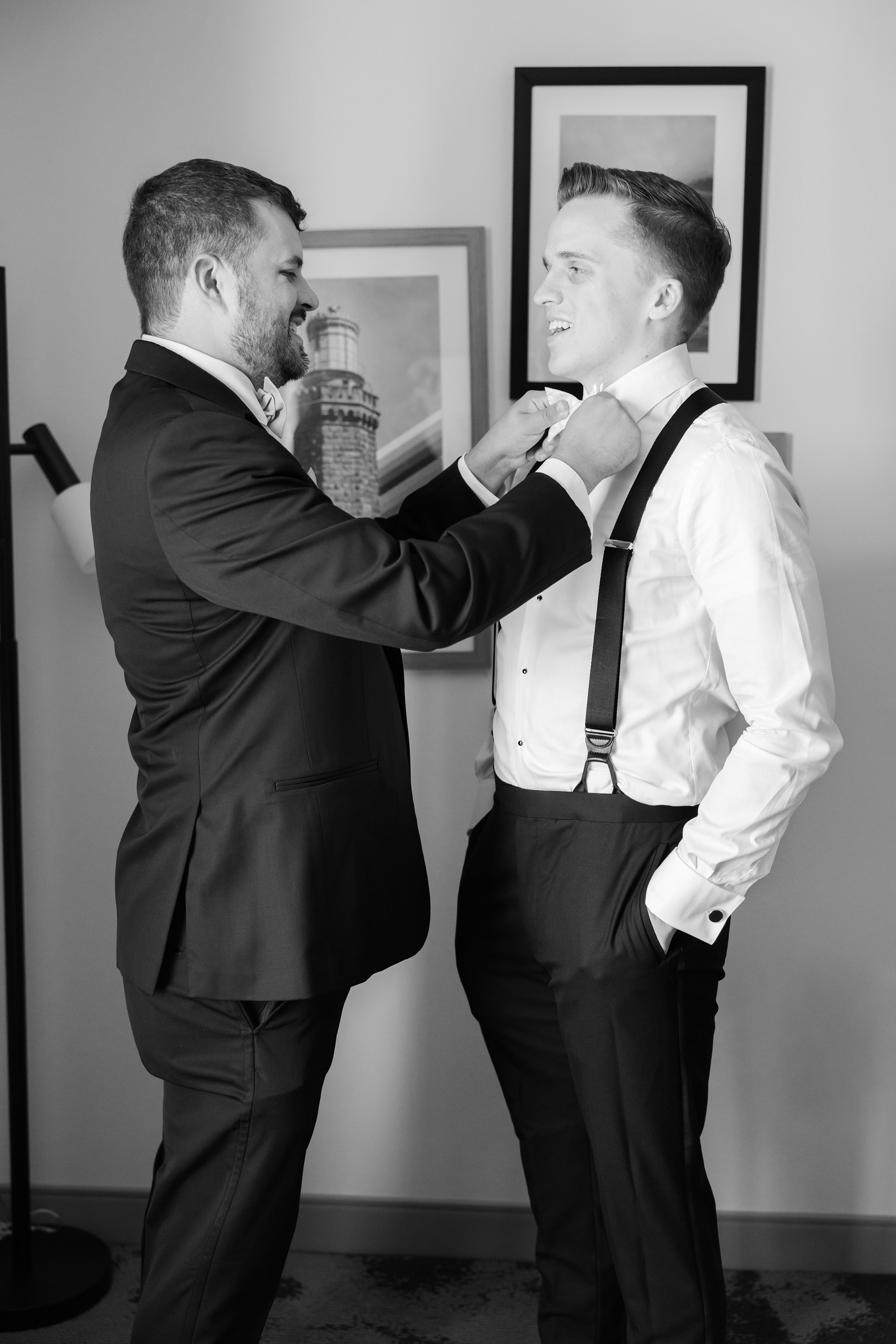 Groomsman helps the groom adjust his bow tie while both smile during preparation.