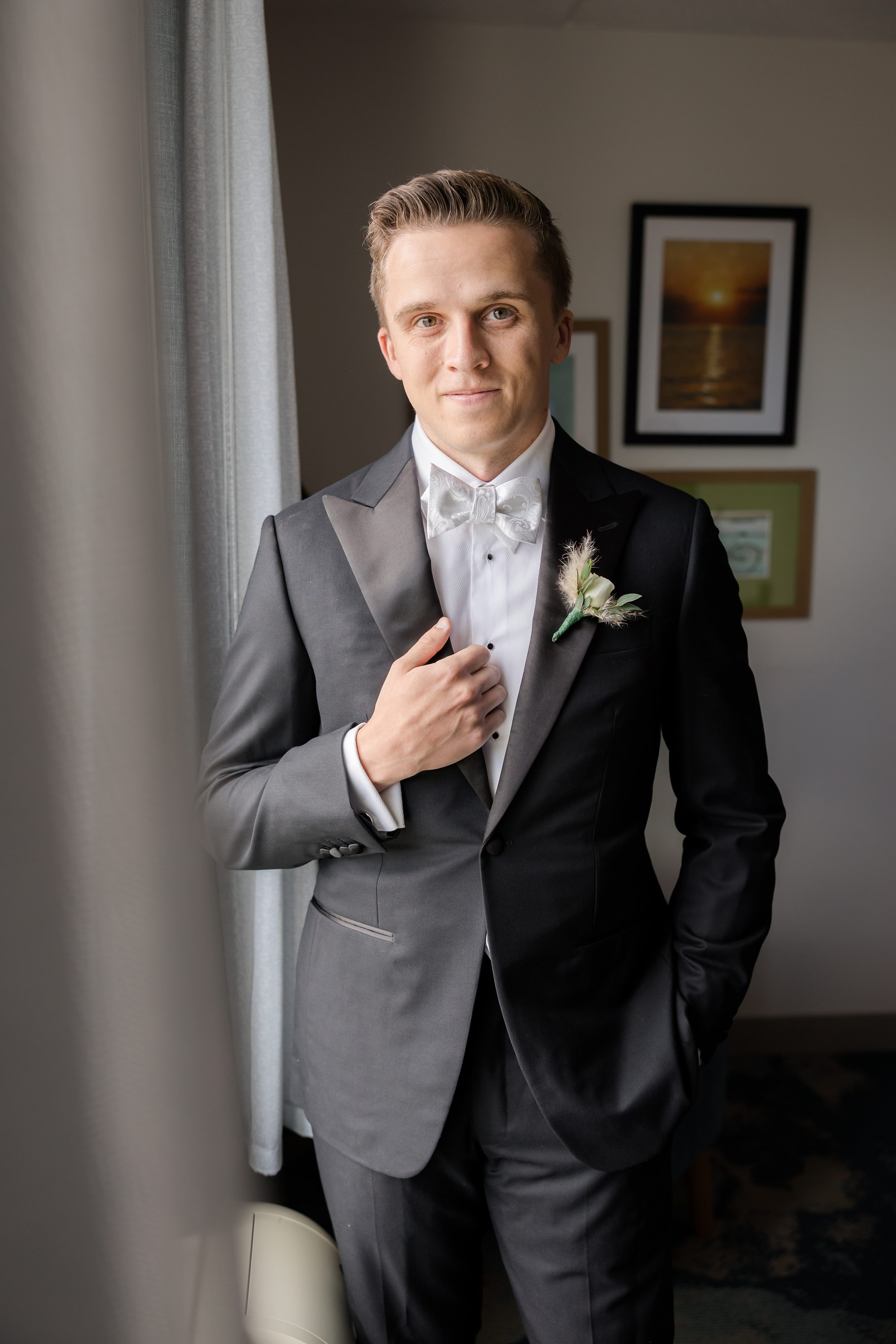Groom in a black tuxedo adjusts his jacket while standing near a window with soft natural light.