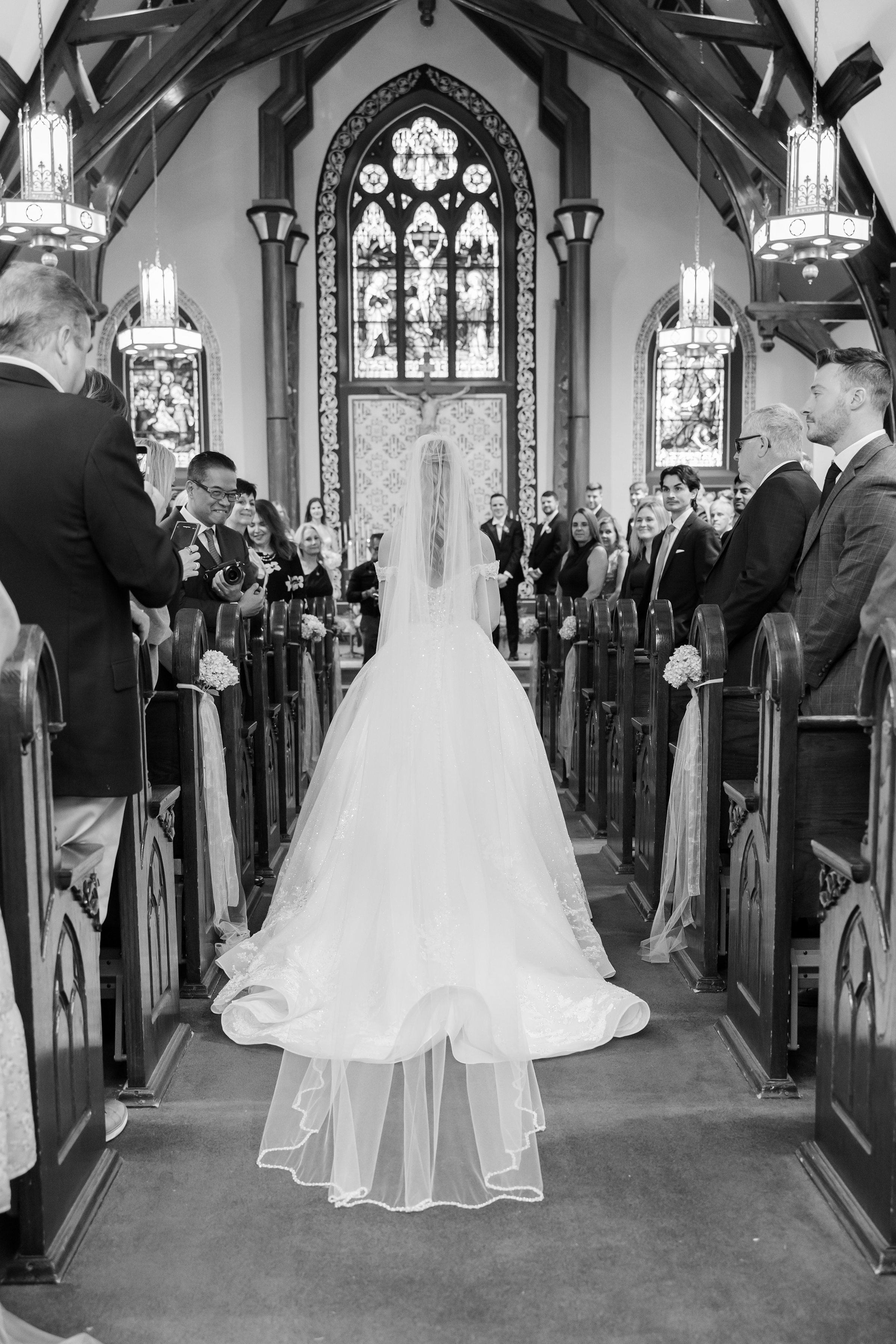 Bride walks down the church aisle toward the groom, viewed from behind with guests seated on both sides.