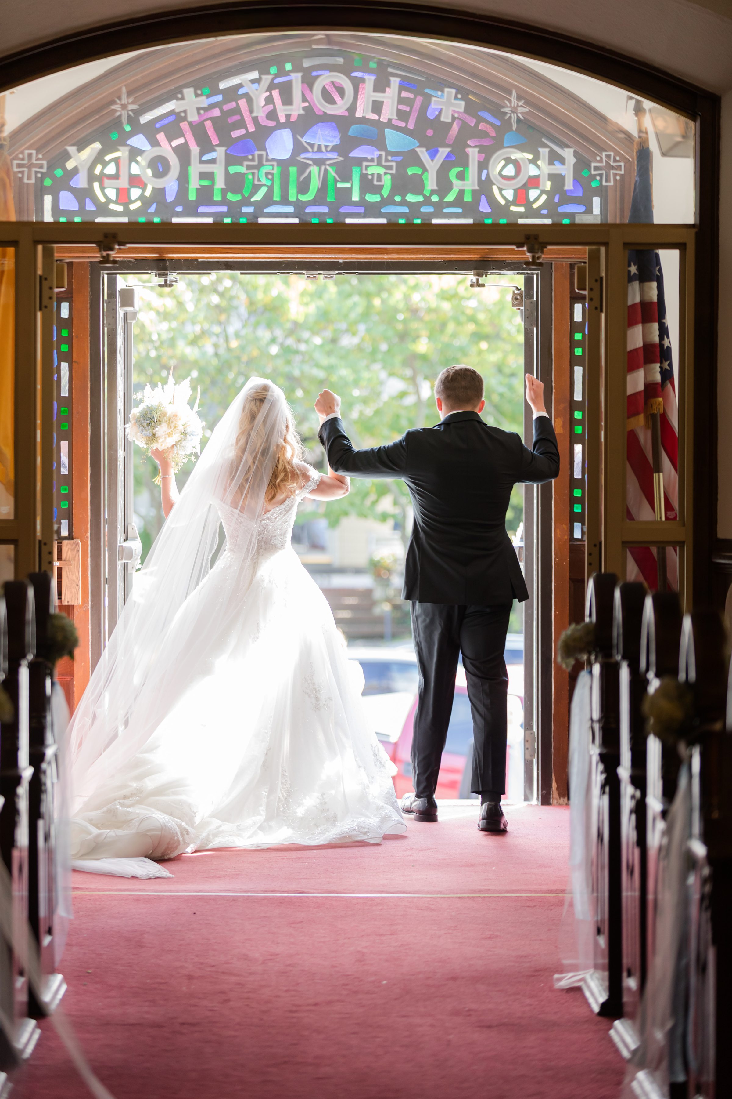 Bride and groom walk out of the church doorway from behind, raising their arms in celebration.