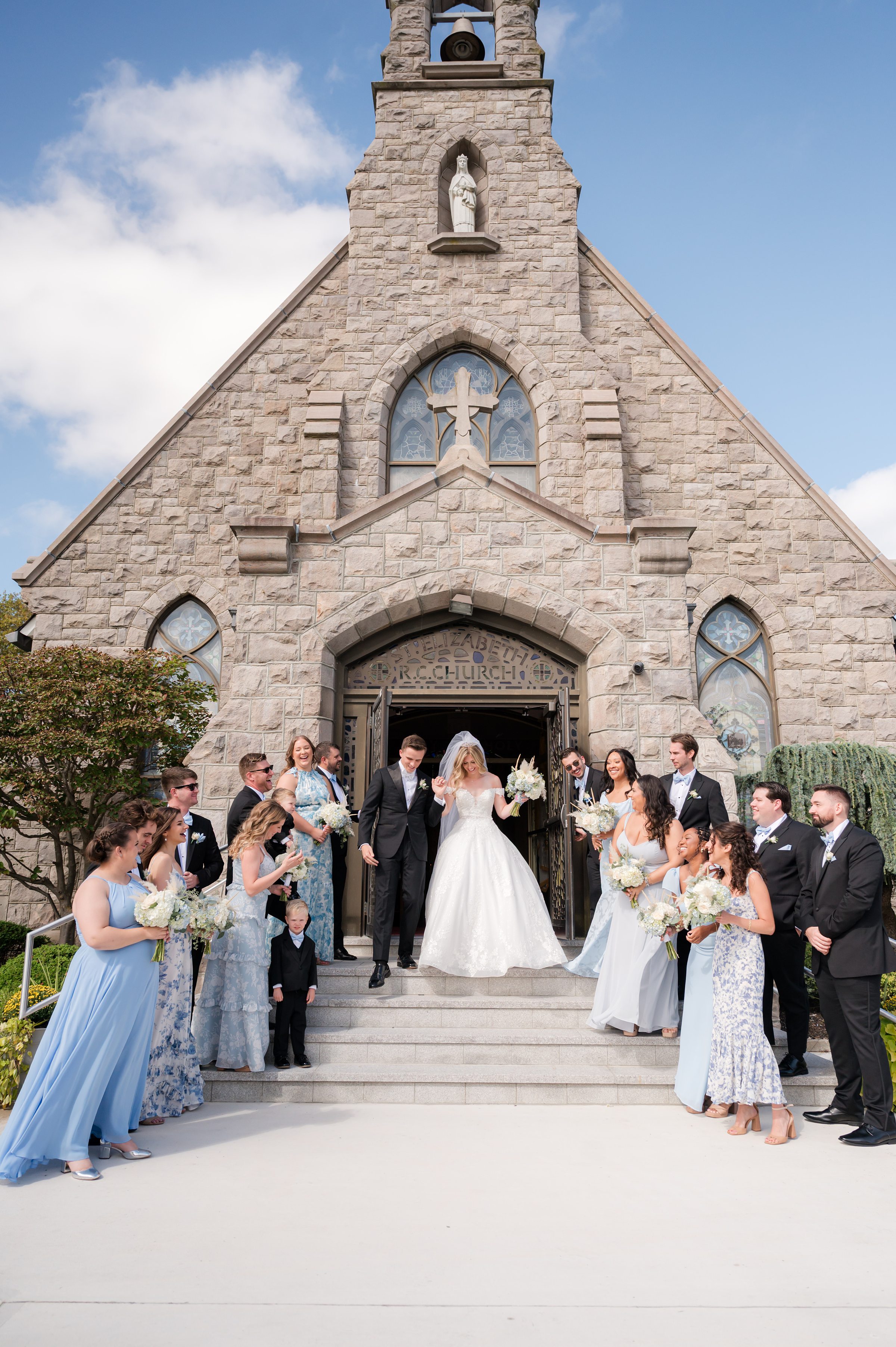 Bride and groom walk down church steps surrounded by bridesmaids and groomsmen holding bouquets.