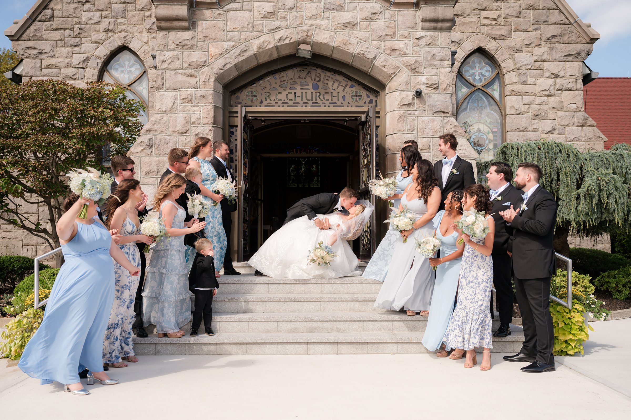 Bride and groom share a dramatic dip kiss on church steps as wedding party members stand on either side cheering.
