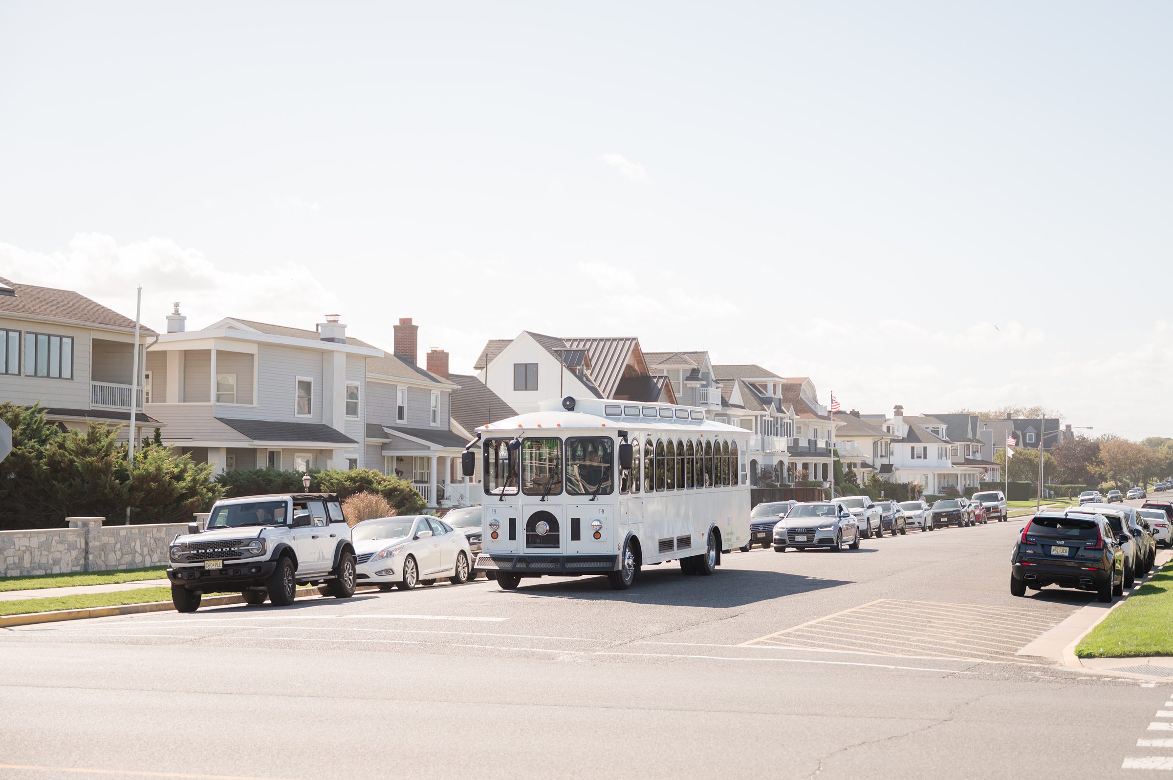 White trolley drives down a residential street lined with beach style houses and parked cars.