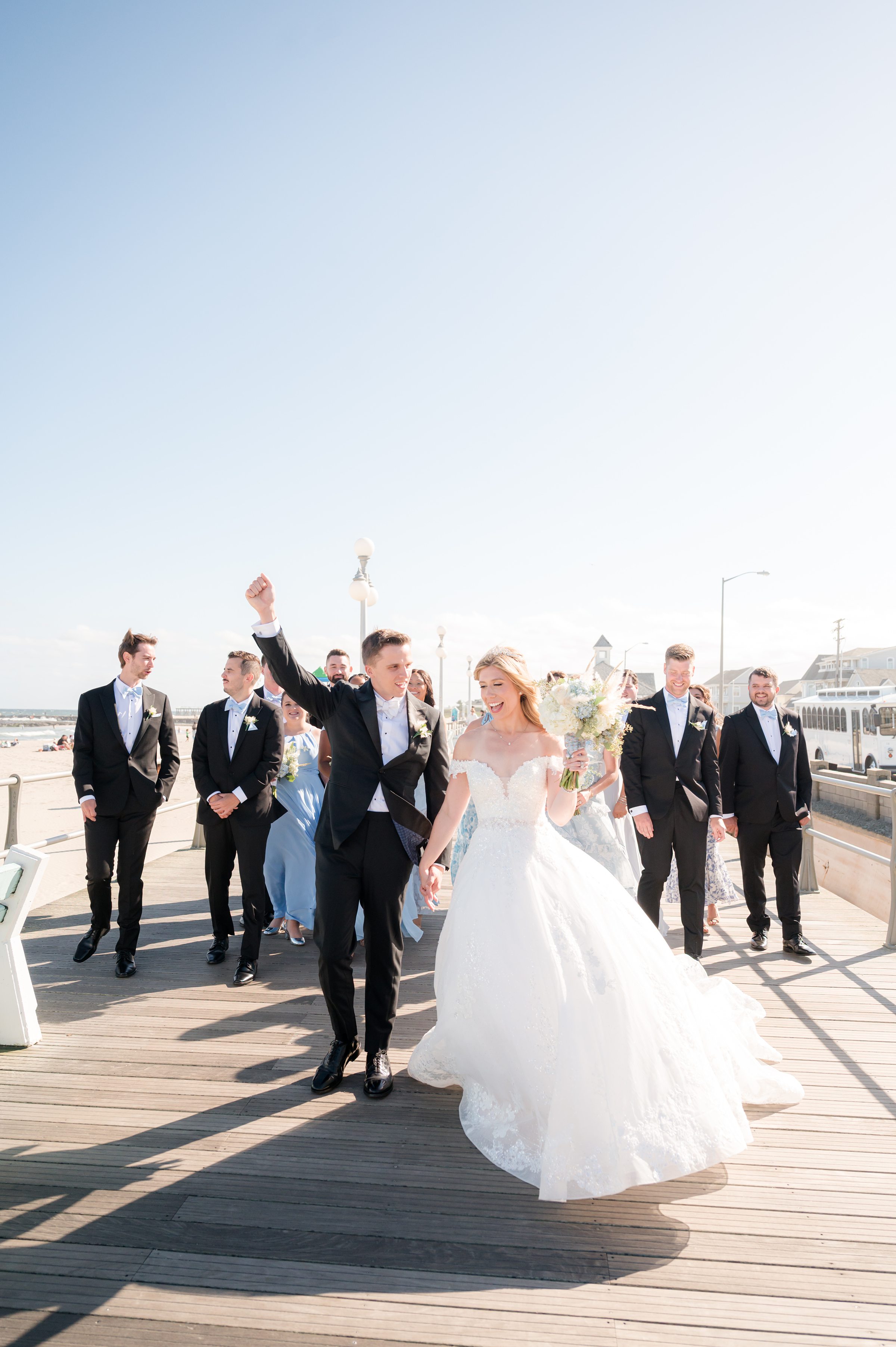 Bride and groom lead their wedding party walking down a boardwalk, smiling and celebrating together.