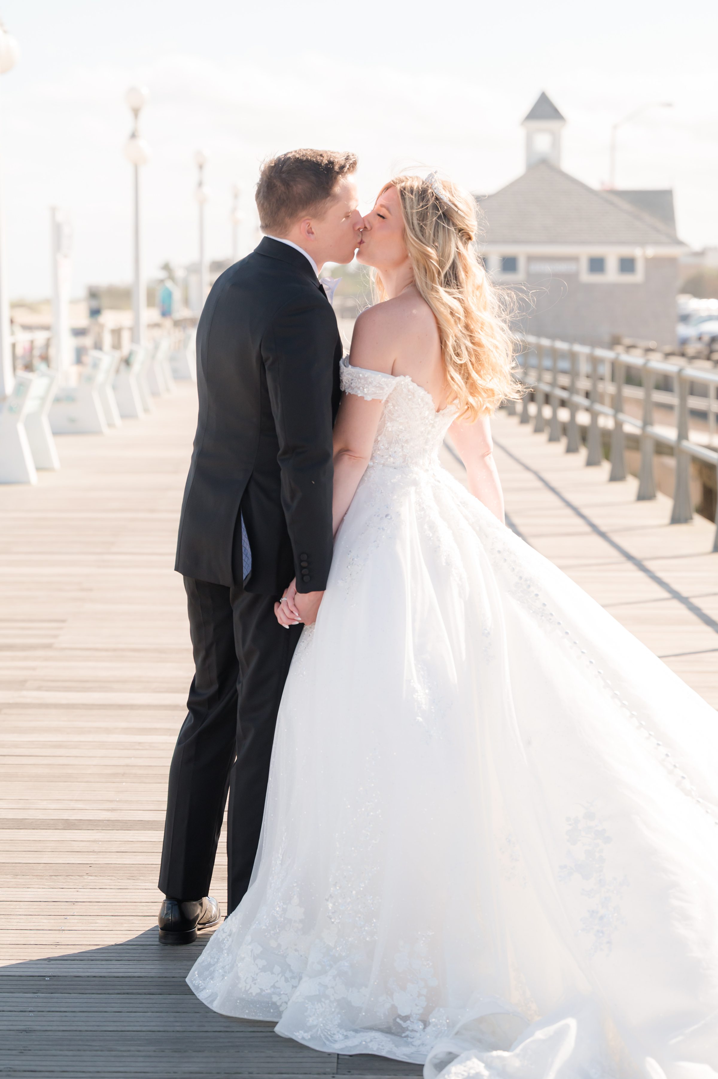 Bride and groom kiss while holding hands on a sunlit boardwalk, with soft beach scenery behind them.
