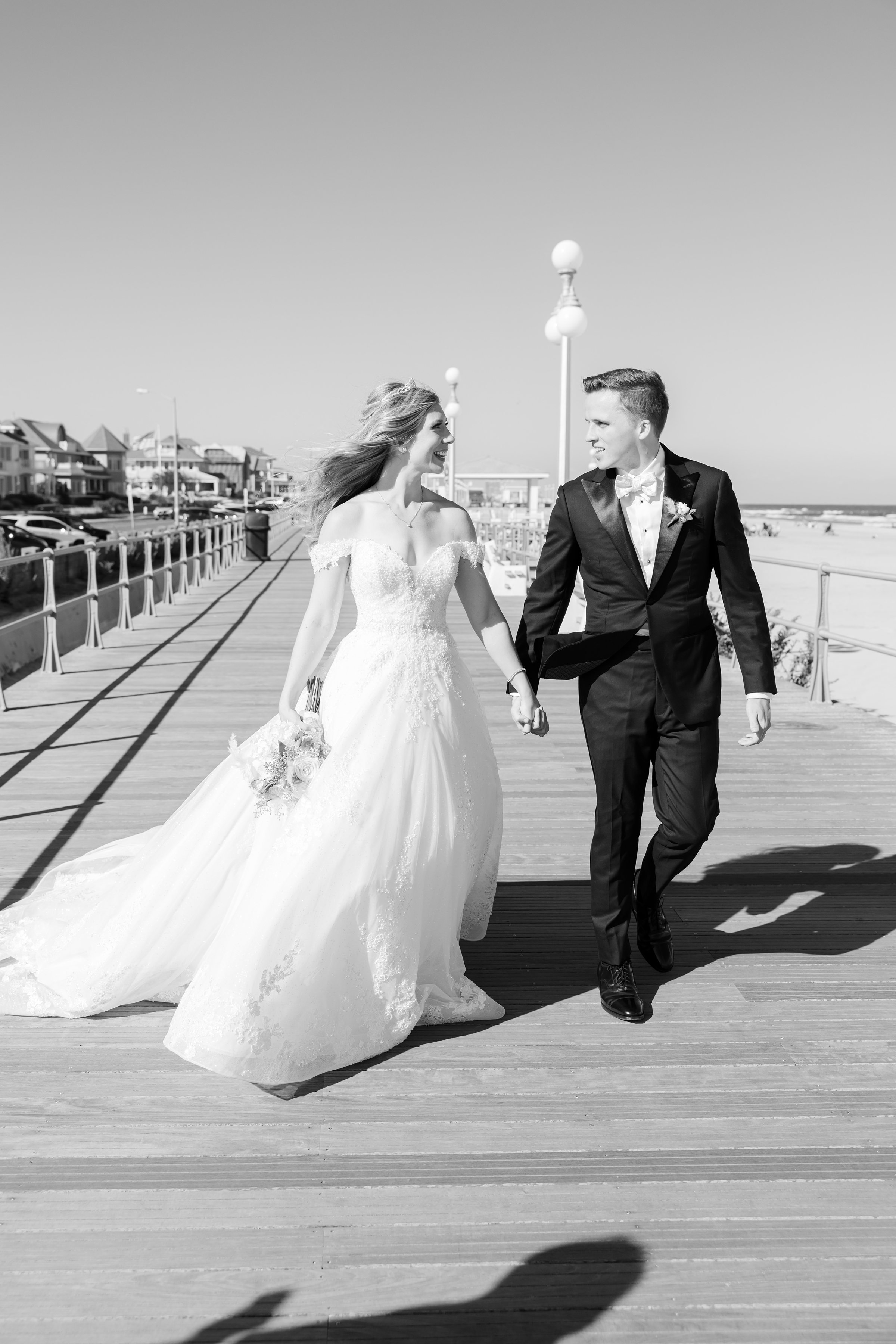 Bride and groom walk together along a beachside boardwalk, smiling at each other with ocean in the background.