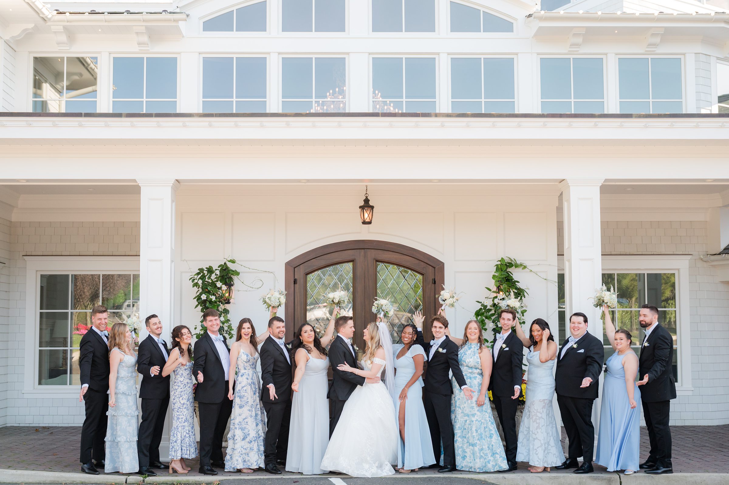 Bride and groom kiss in the center while bridesmaids and groomsmen cheer with raised bouquets and arms outside a large white building.