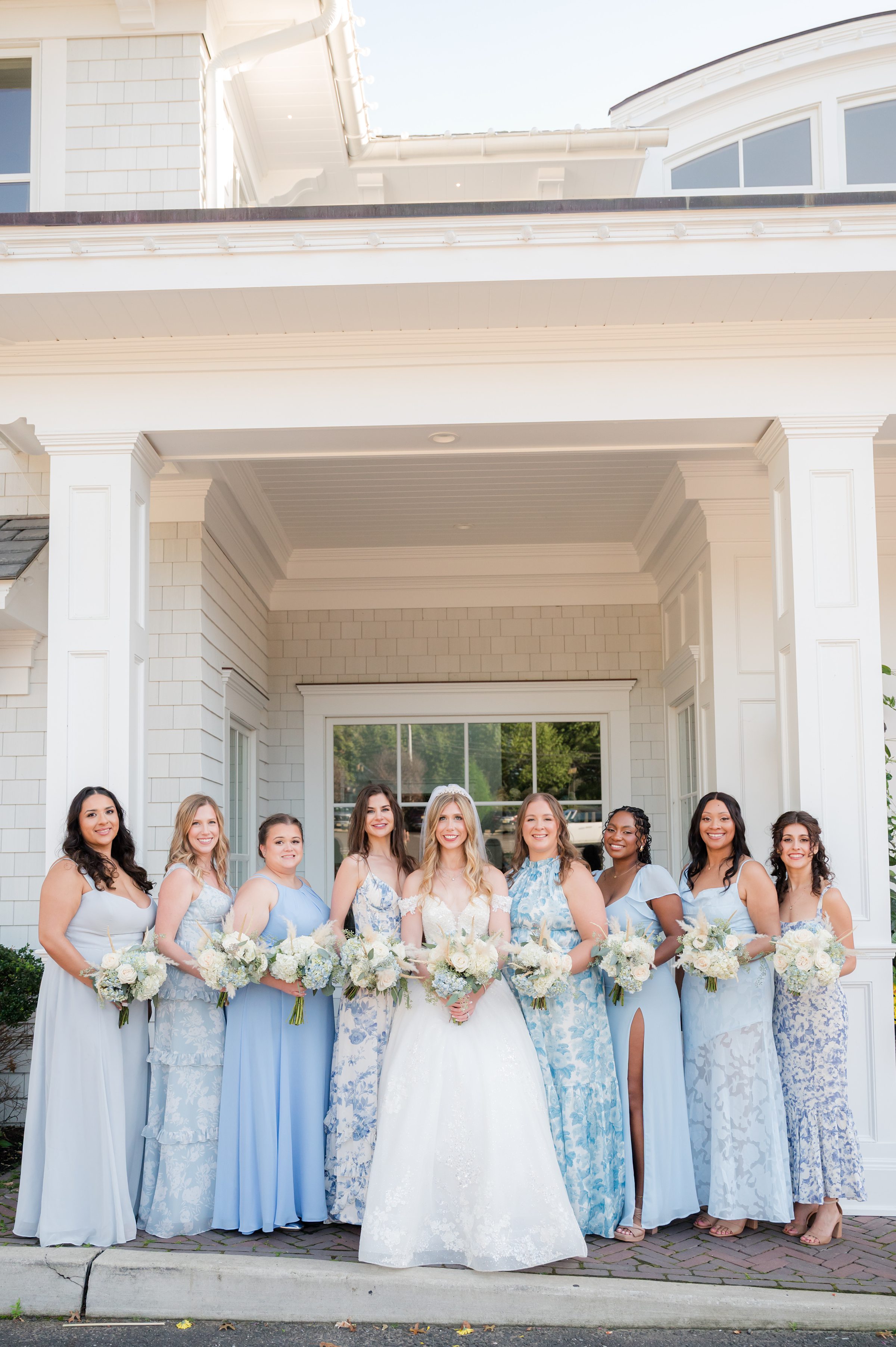 Bride in a white lace gown stands in the center surrounded by bridesmaids in light blue dresses holding bouquets, posed in front of a white building.