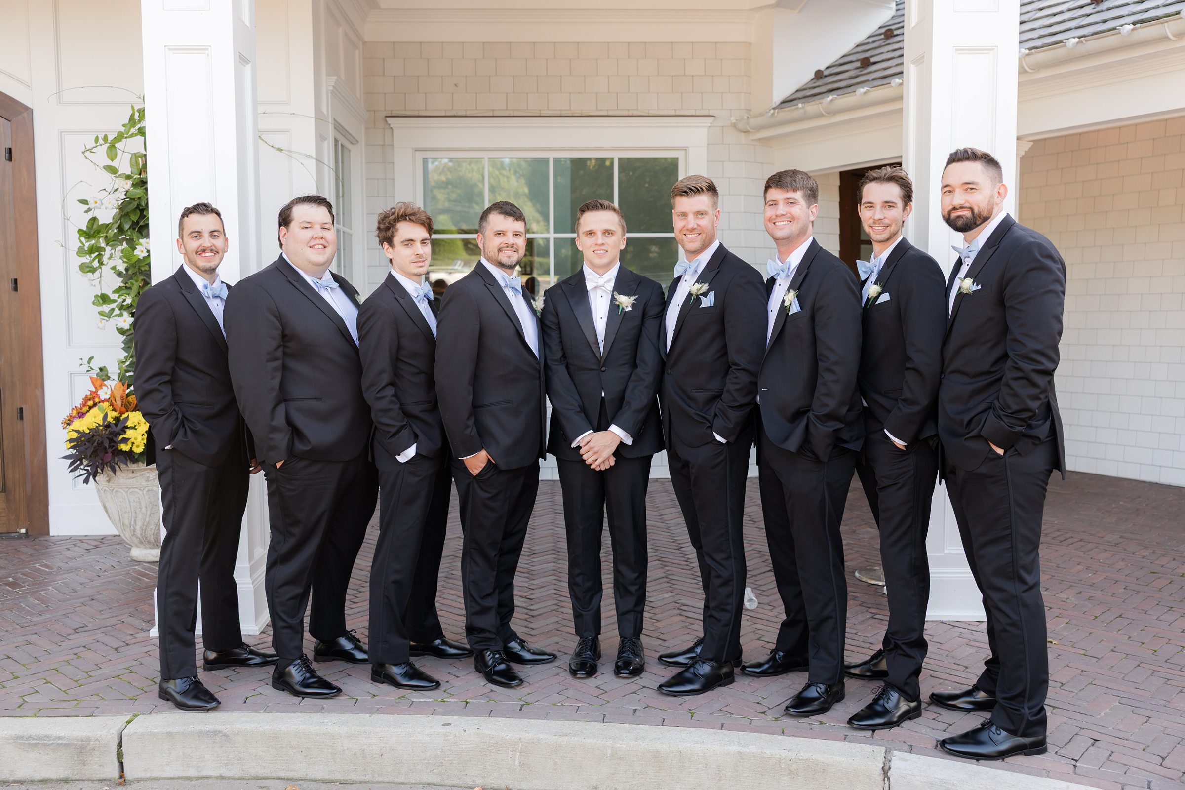 Groom stands centered with groomsmen in black tuxedos and bow ties, posing outside a white building entrance on a sunny day.