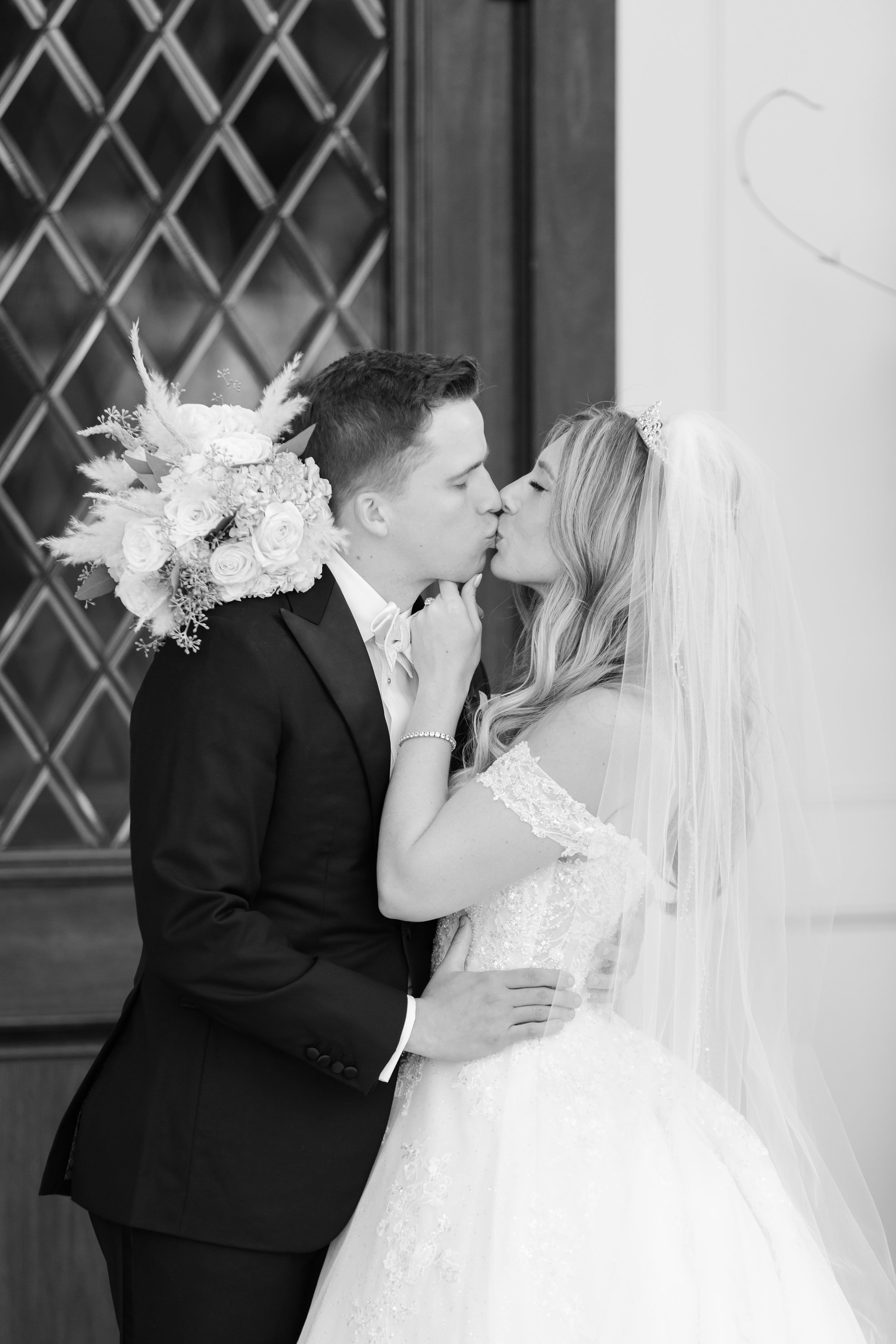 Bride and groom sharing a gentle kiss, the bride’s veil softly draped around them as she holds her bouquet, creating an intimate and romantic moment.
