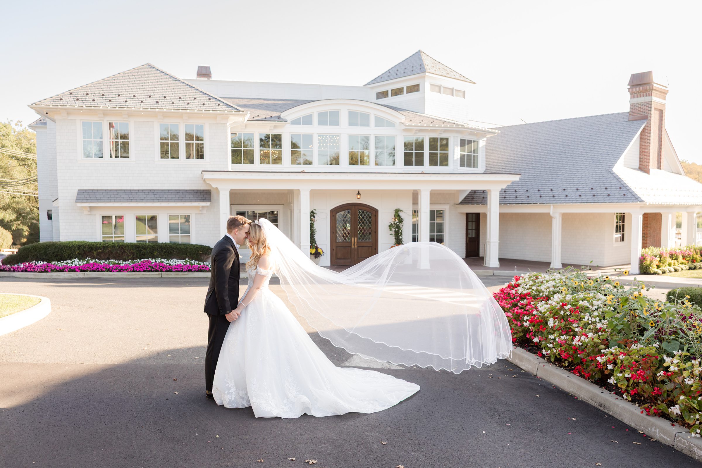 Bride and groom standing outside a charming white venue, her veil flowing like a dream behind them.