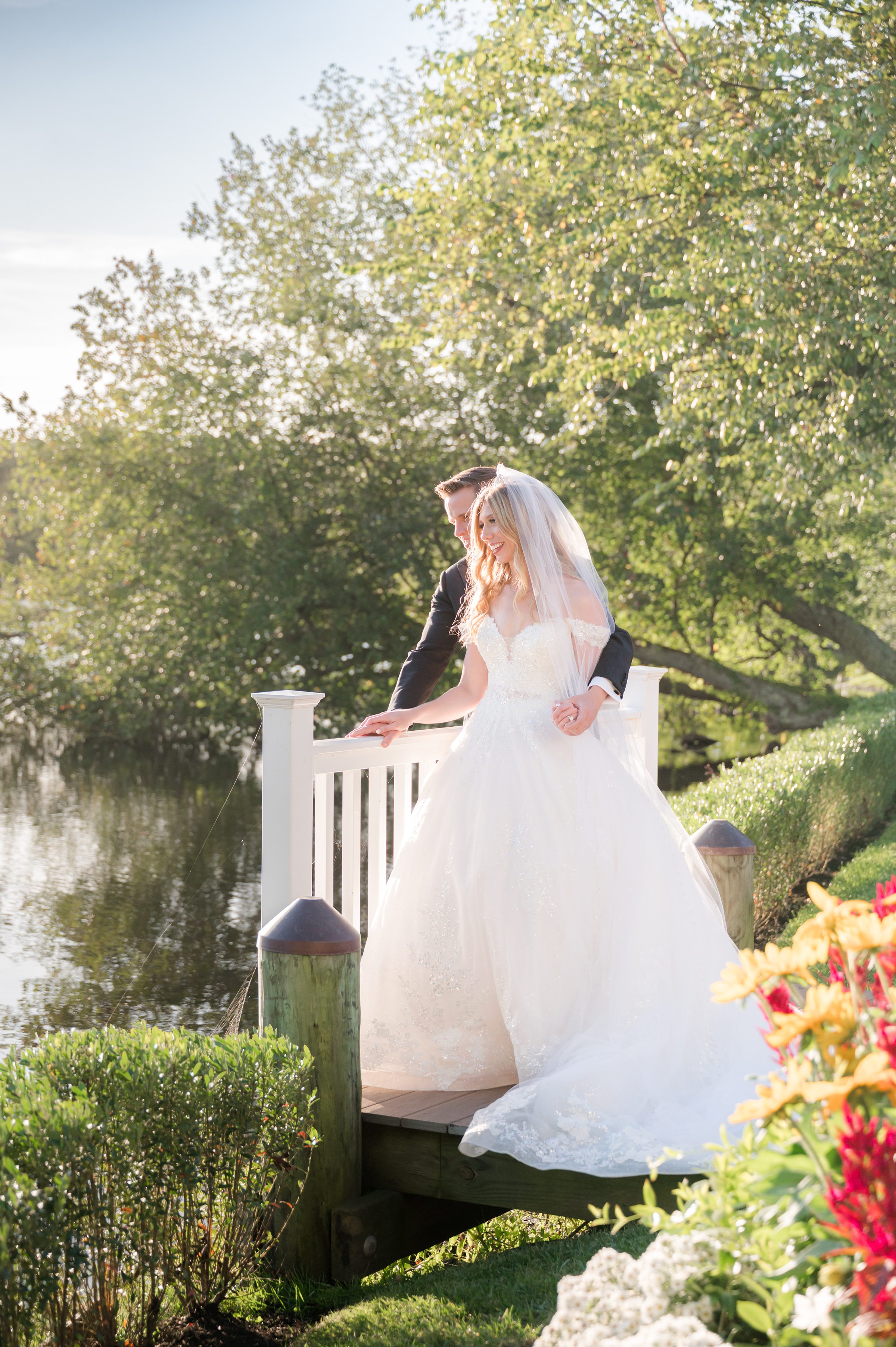 Bride and groom standing together on a sunlit dock by the water, surrounded by lush greenery, sharing a peaceful and romantic moment in soft golden light.
