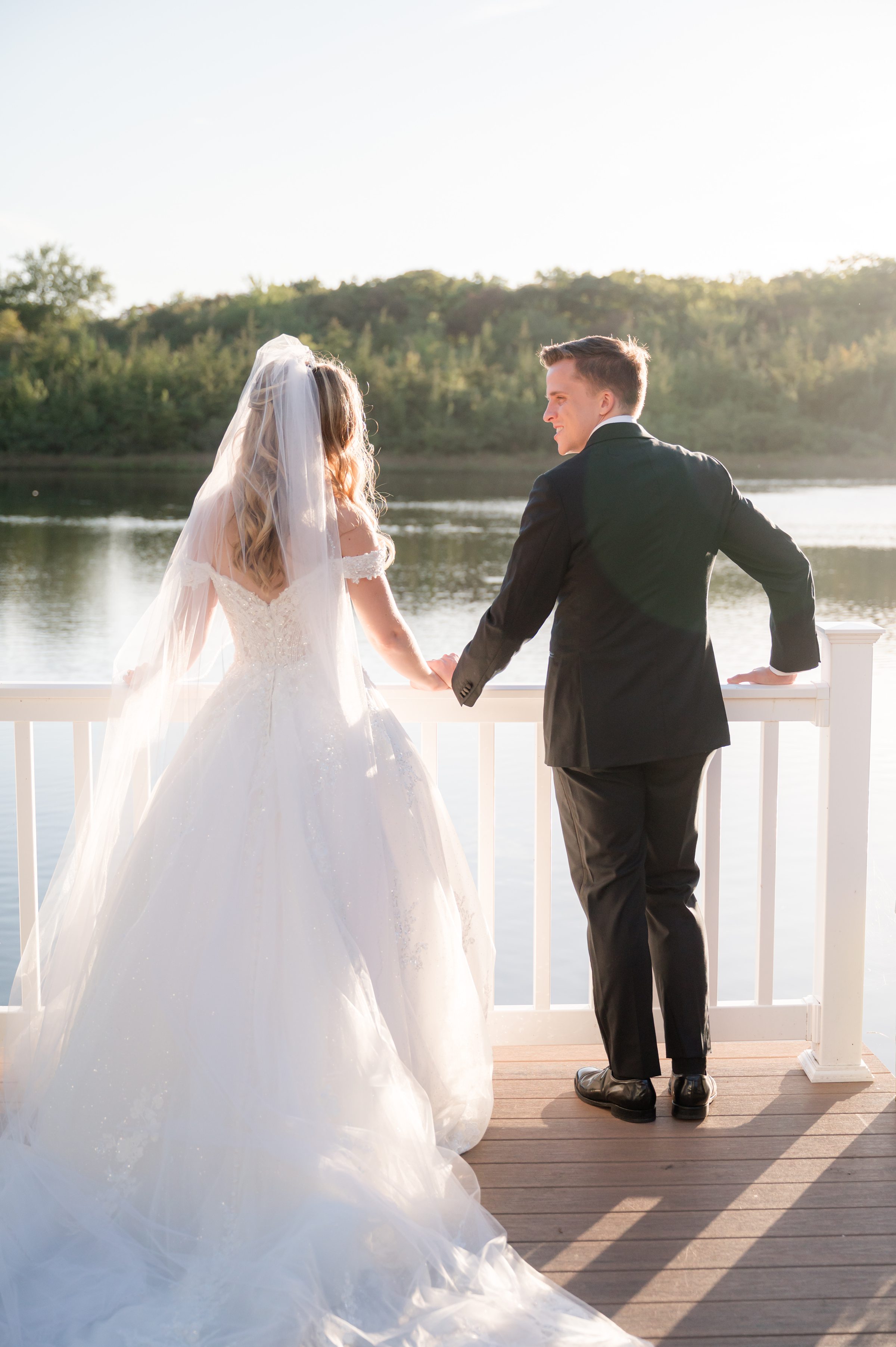 Bride and groom holding hands at a waterfront railing, bathed in warm golden sunlight as they look out over the water, sharing a quiet and romantic moment together.