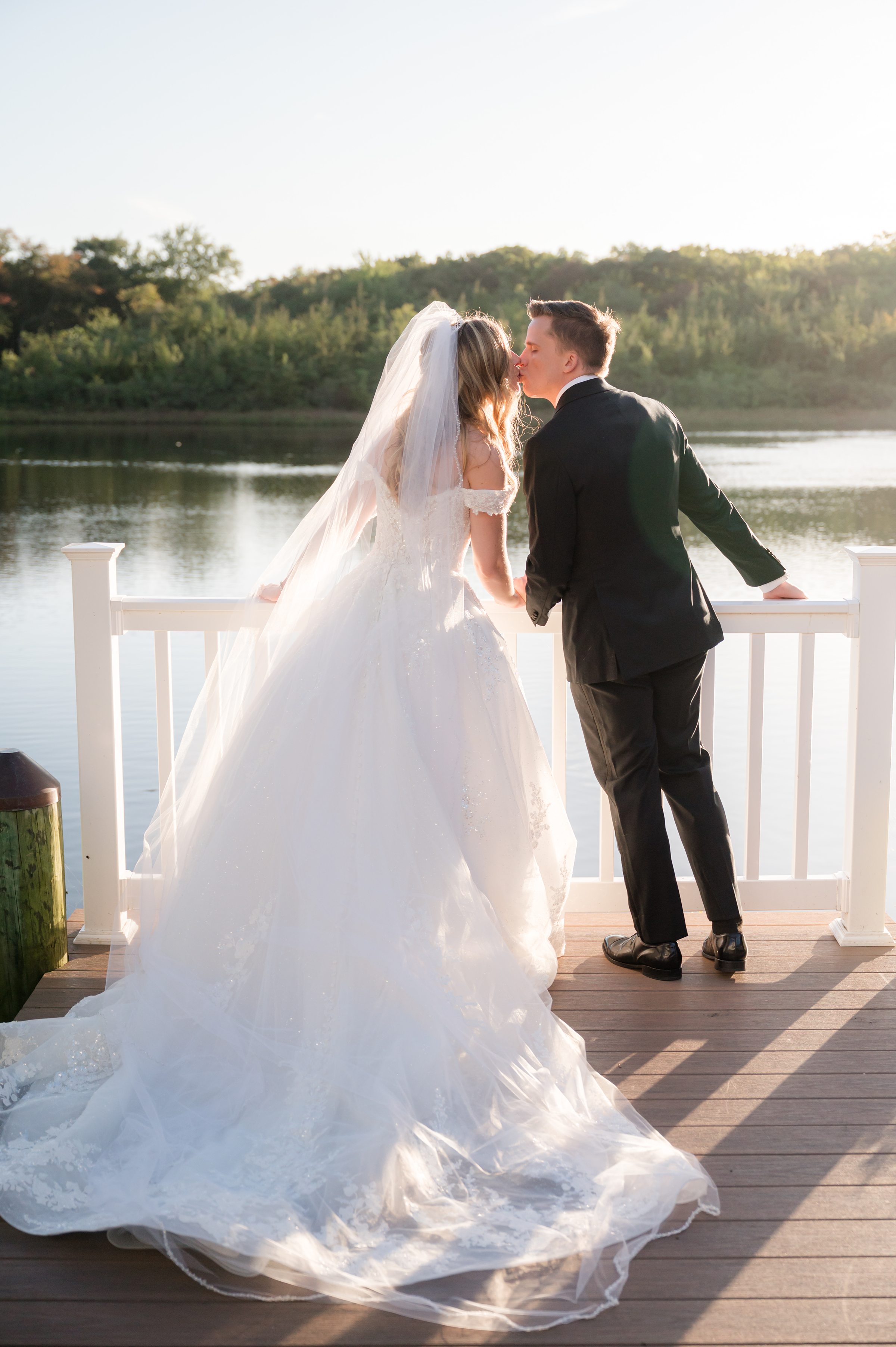 Bride and groom sharing a tender sunset kiss by the water, wrapped in golden light.