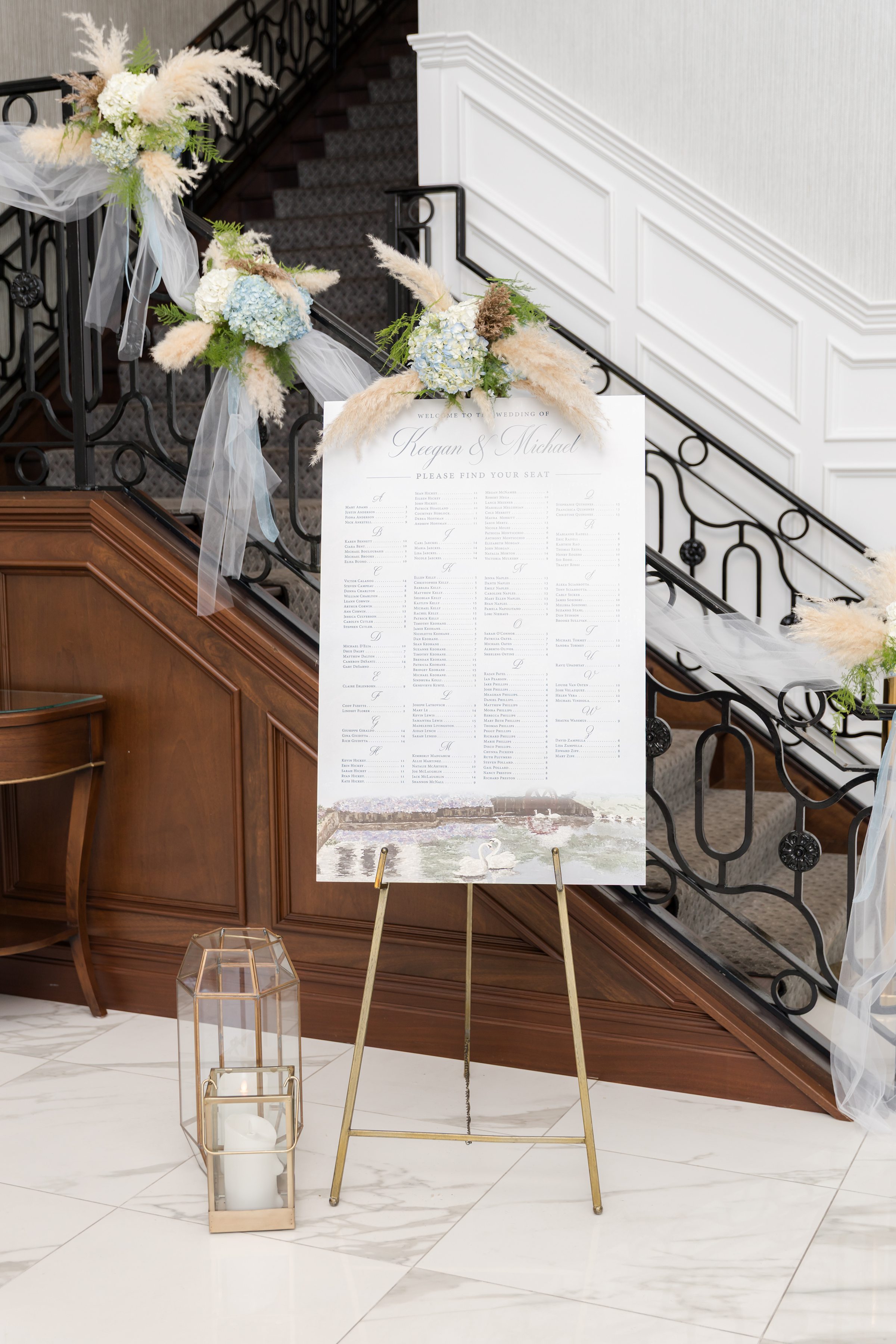 Romantic seating chart display framed by airy florals and flowing fabric along a grand staircase.