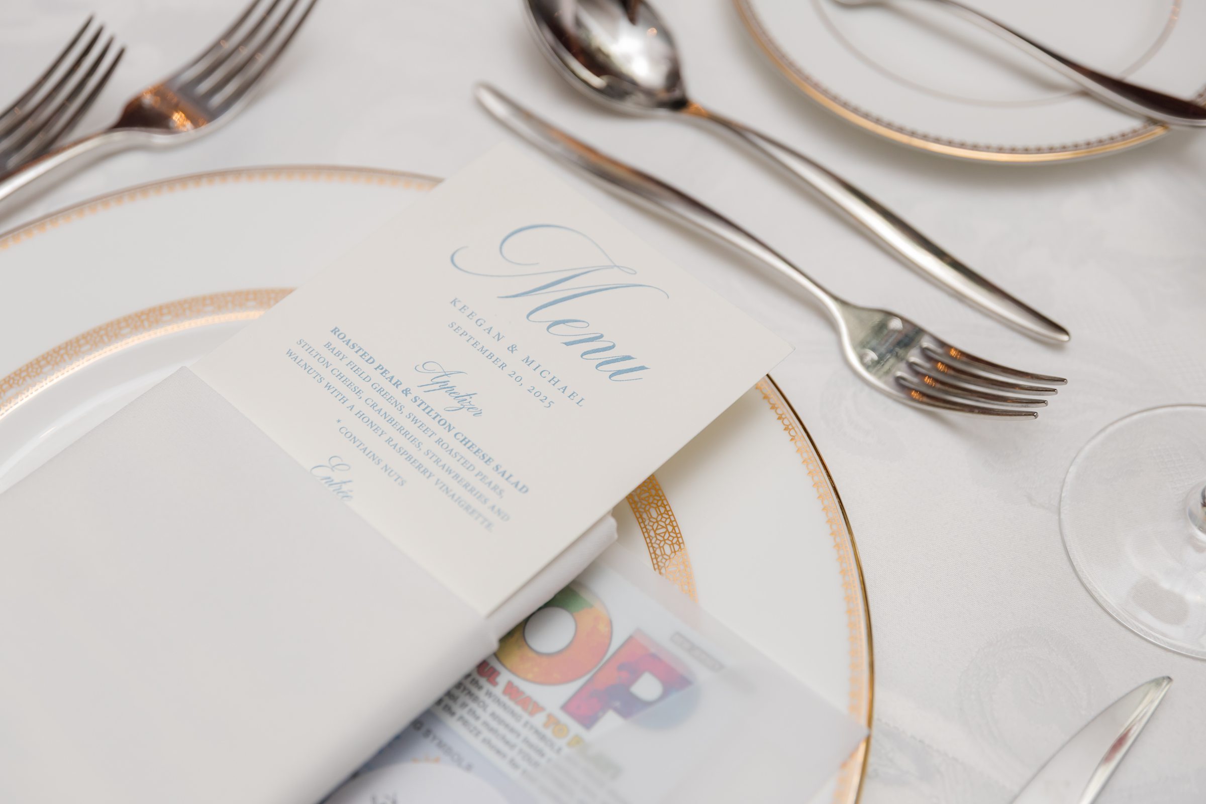Elegant wedding place setting with gold rimmed neatly arranged silverware, and a personalized menu card resting on a folded napkin, creating a refined and romantic tables cape.