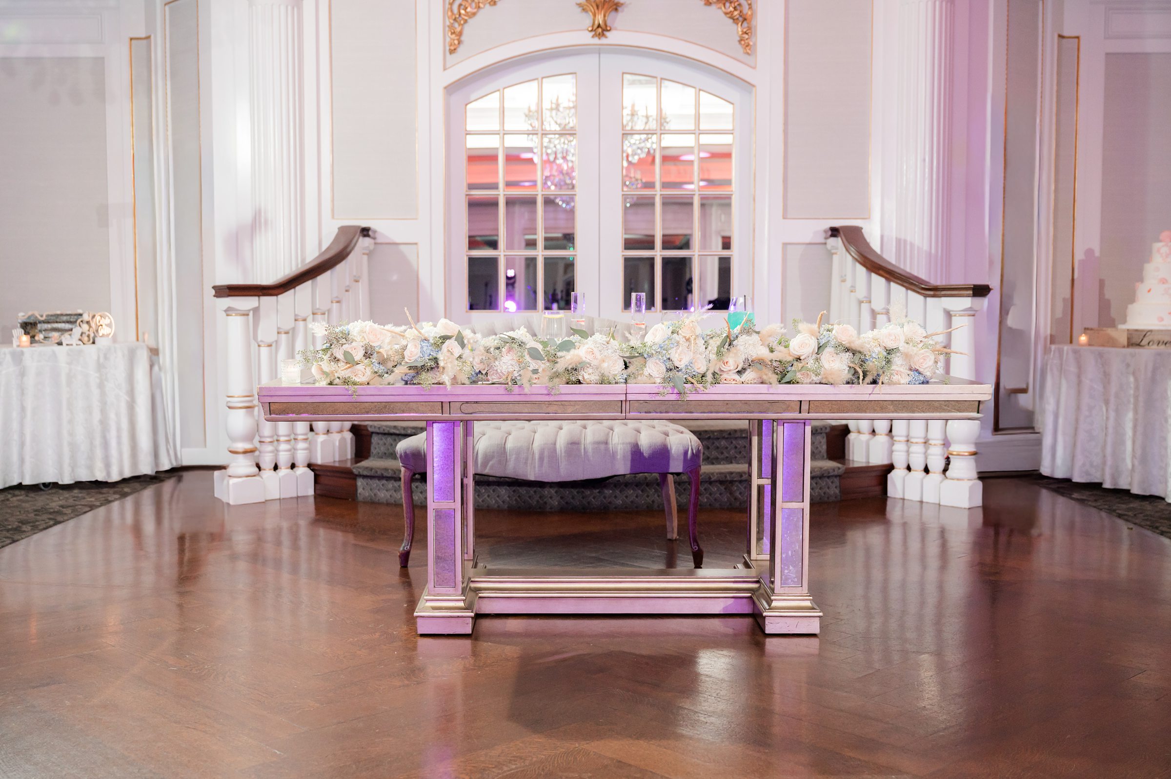 Elegant head table with a long floral arrangement and upholstered bench, set in a bright ballroom with chandeliers, white columns, and soft purple accent lighting.