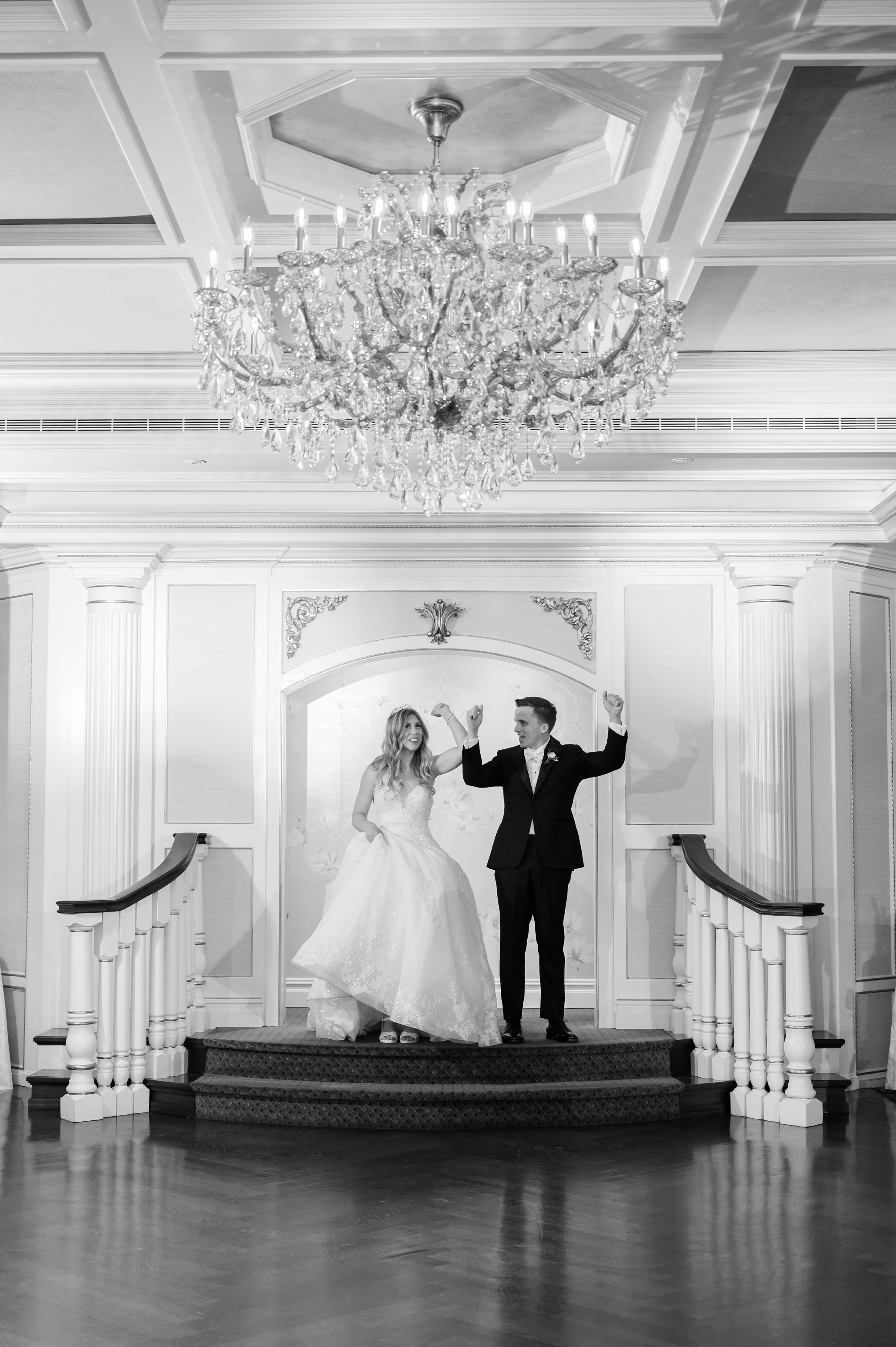 Bride and groom stand at the top of a staircase beneath a large crystal chandelier, raising their hands in celebration in an elegant black and white ballroom setting.