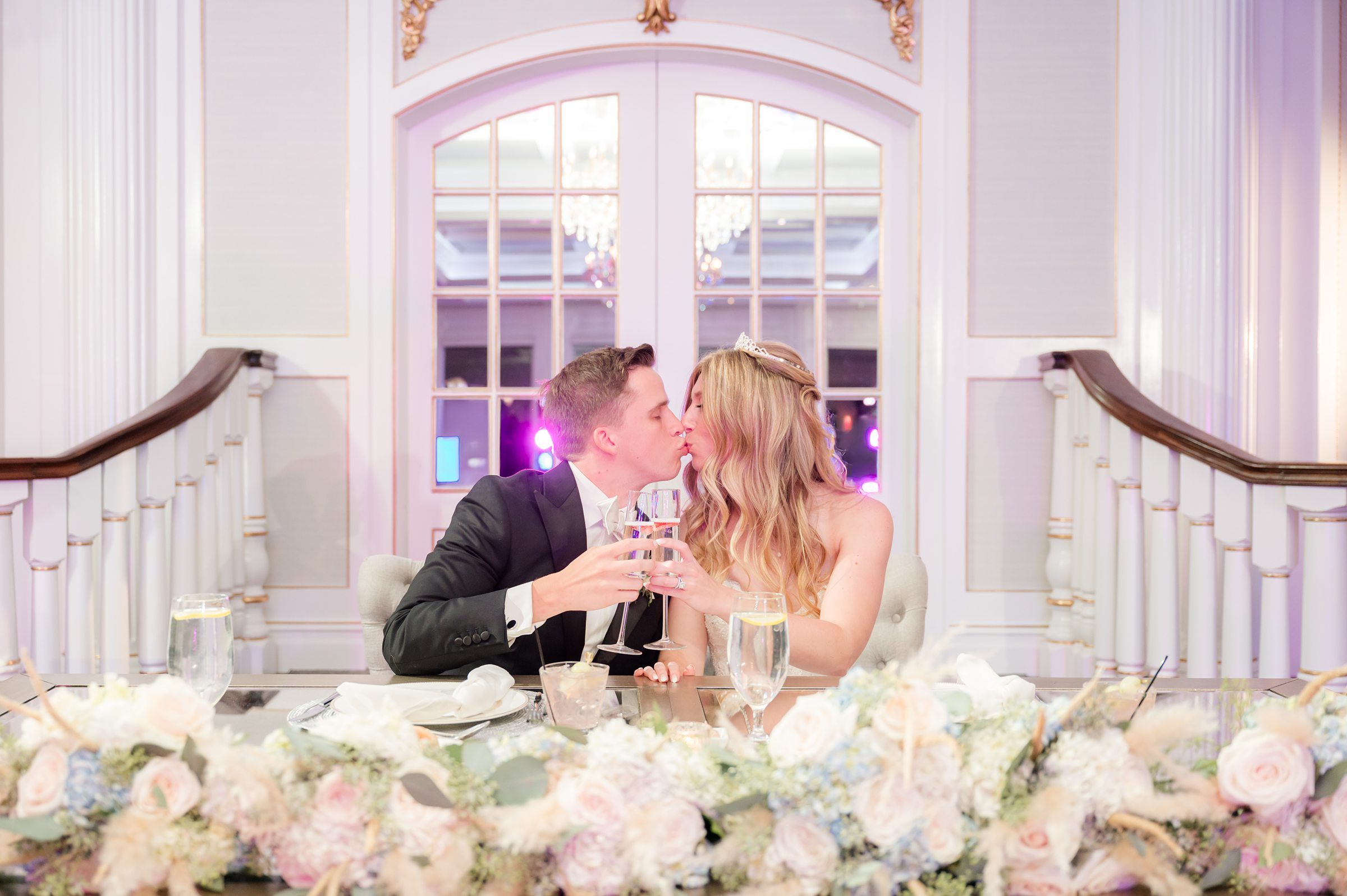 Bride and groom toast with champagne glasses and kiss at their sweetheart table decorated with flowers.