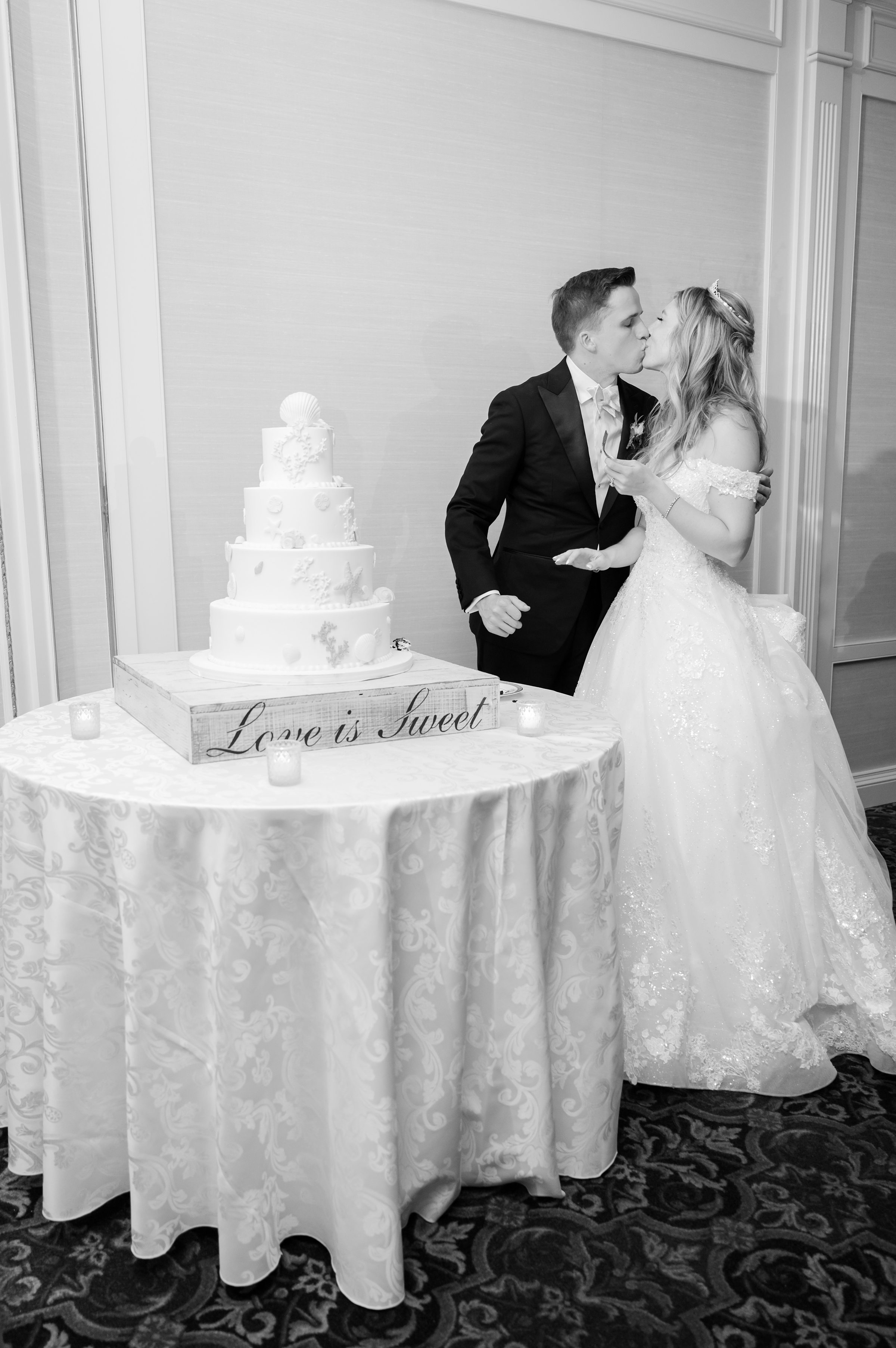 Bride and groom kiss beside a tiered wedding cake on a decorated table.