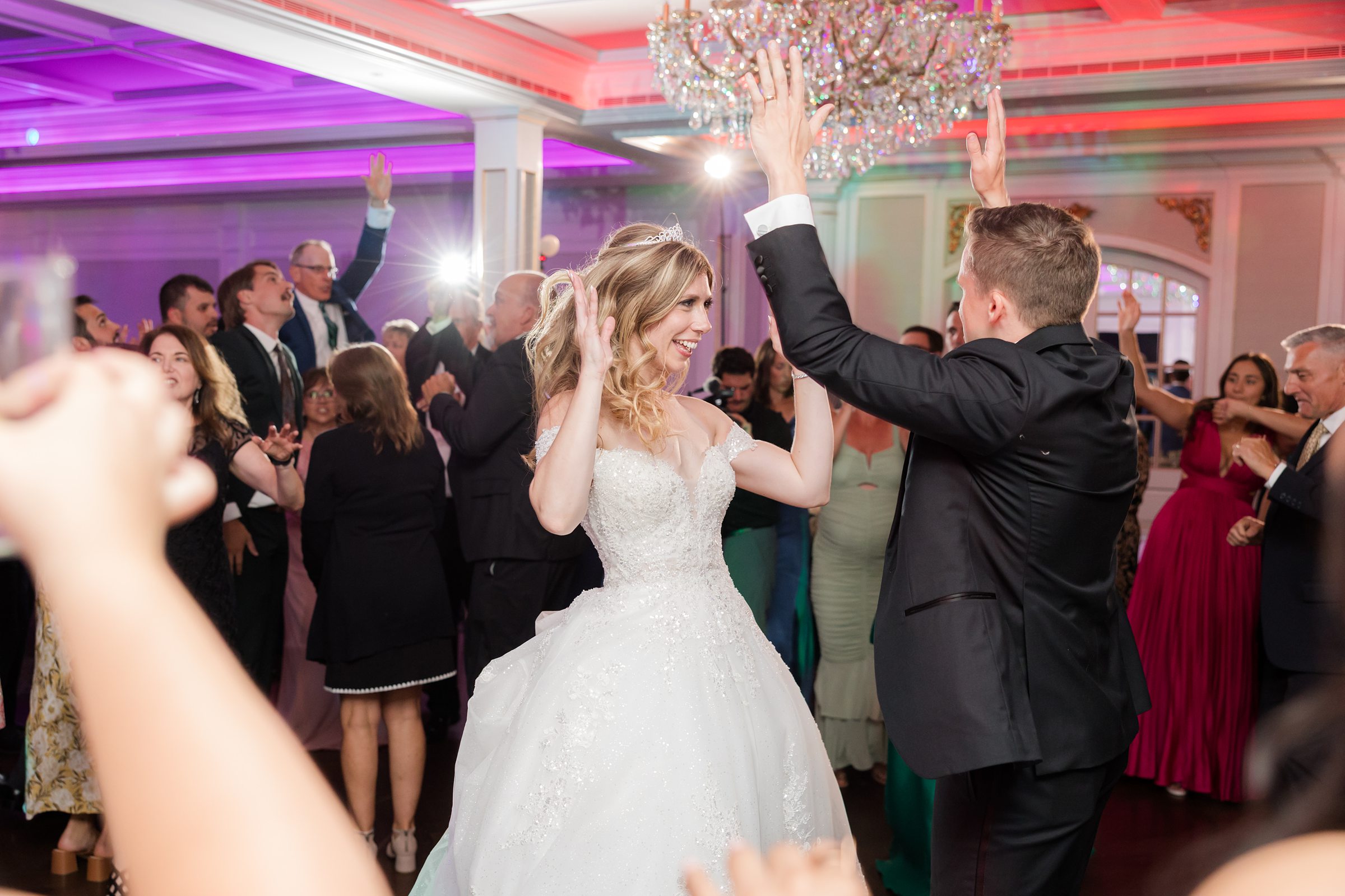 Bride and groom raise their hands mid dance beneath colorful reception lighting and a chandelier.