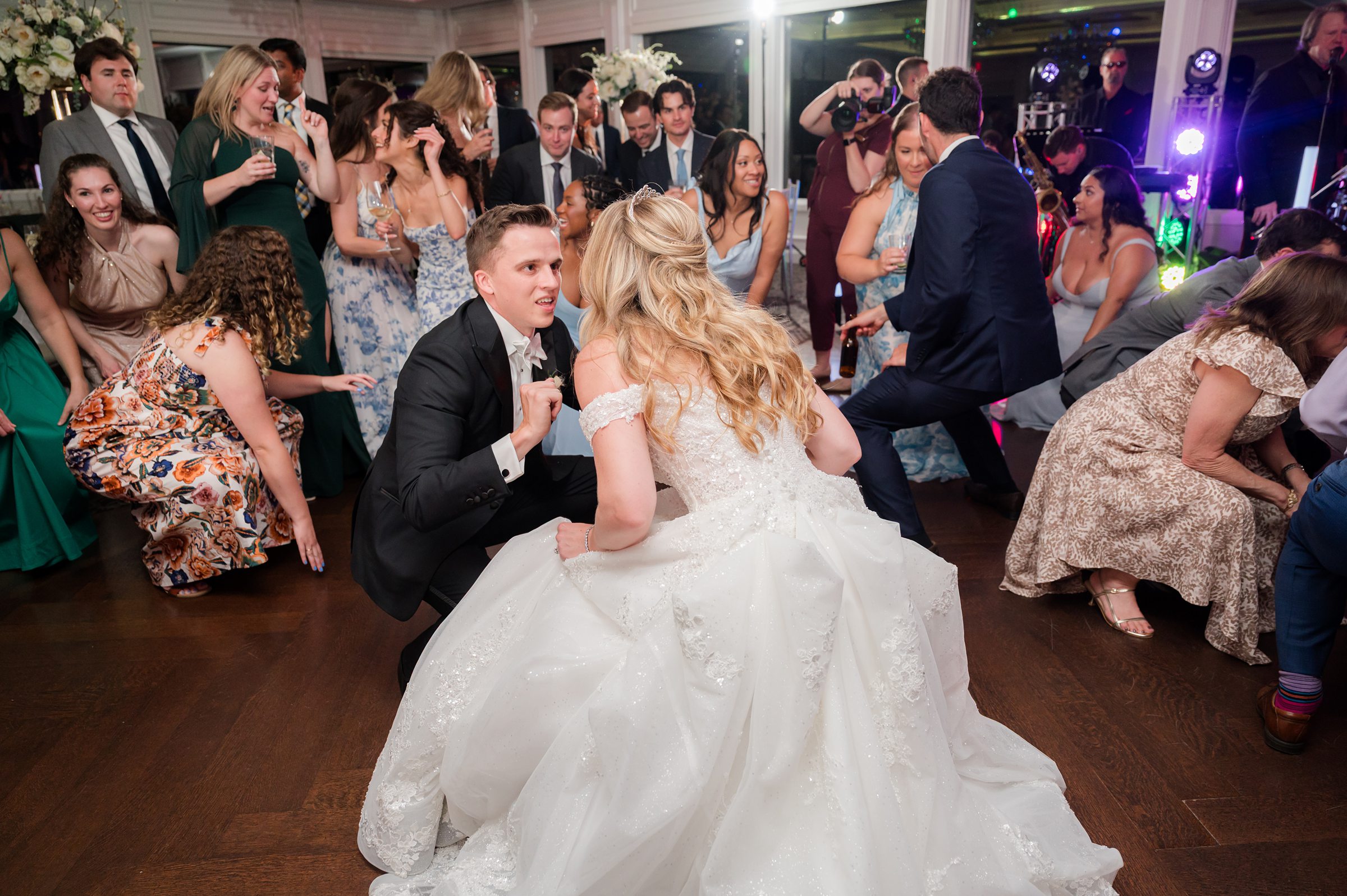 Bride and groom crouch and dance playfully in the center of a lively reception crowd.