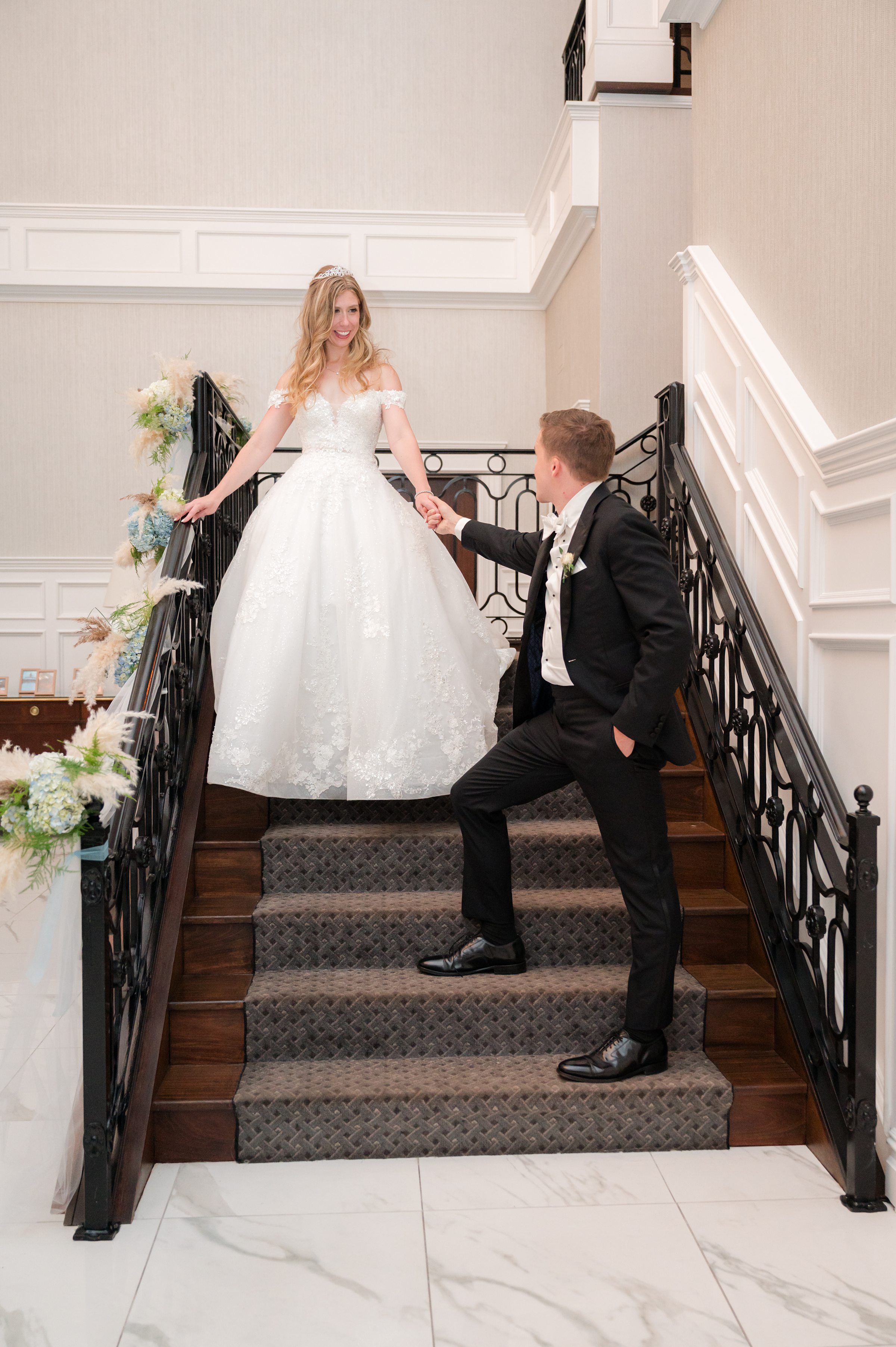 Bride smiles while holding the groom’s hand as she steps down the staircase, her gown flowing.
