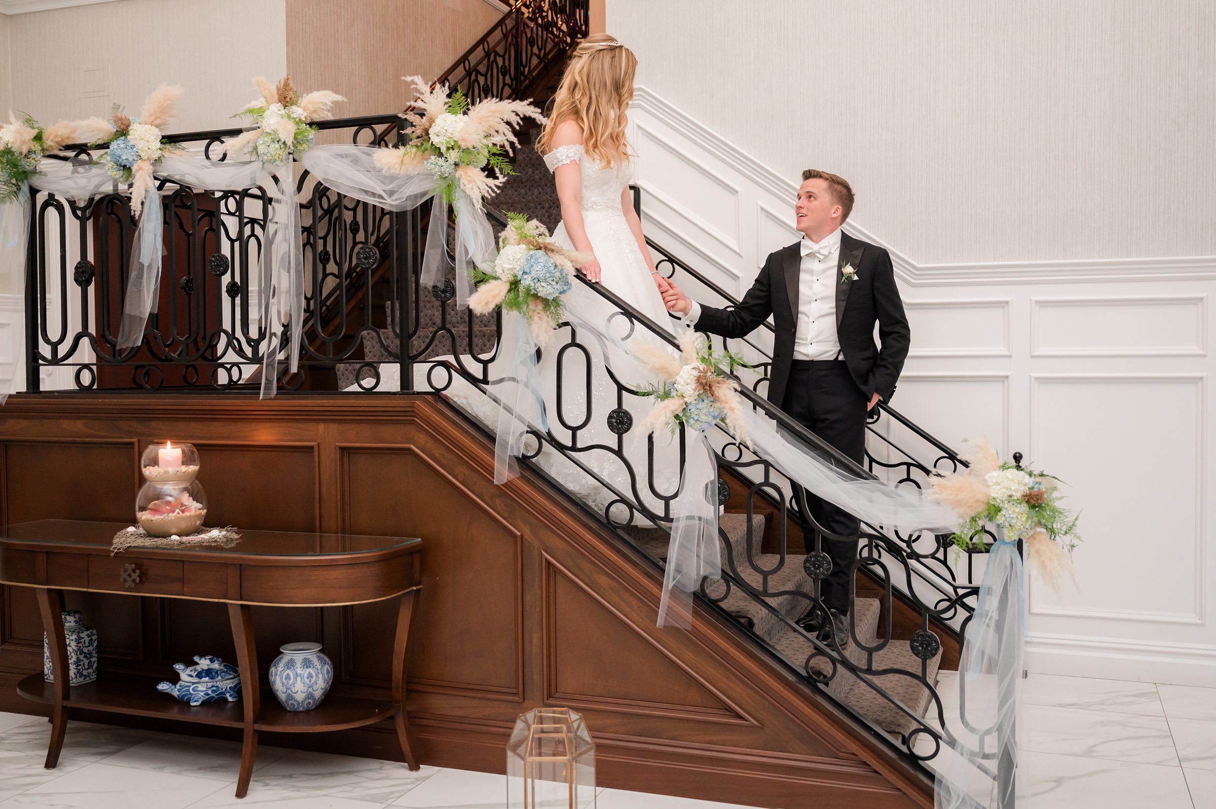 Groom stands below on a staircase holding the bride’s hand as she looks down, with soft floral decorations along the railing.