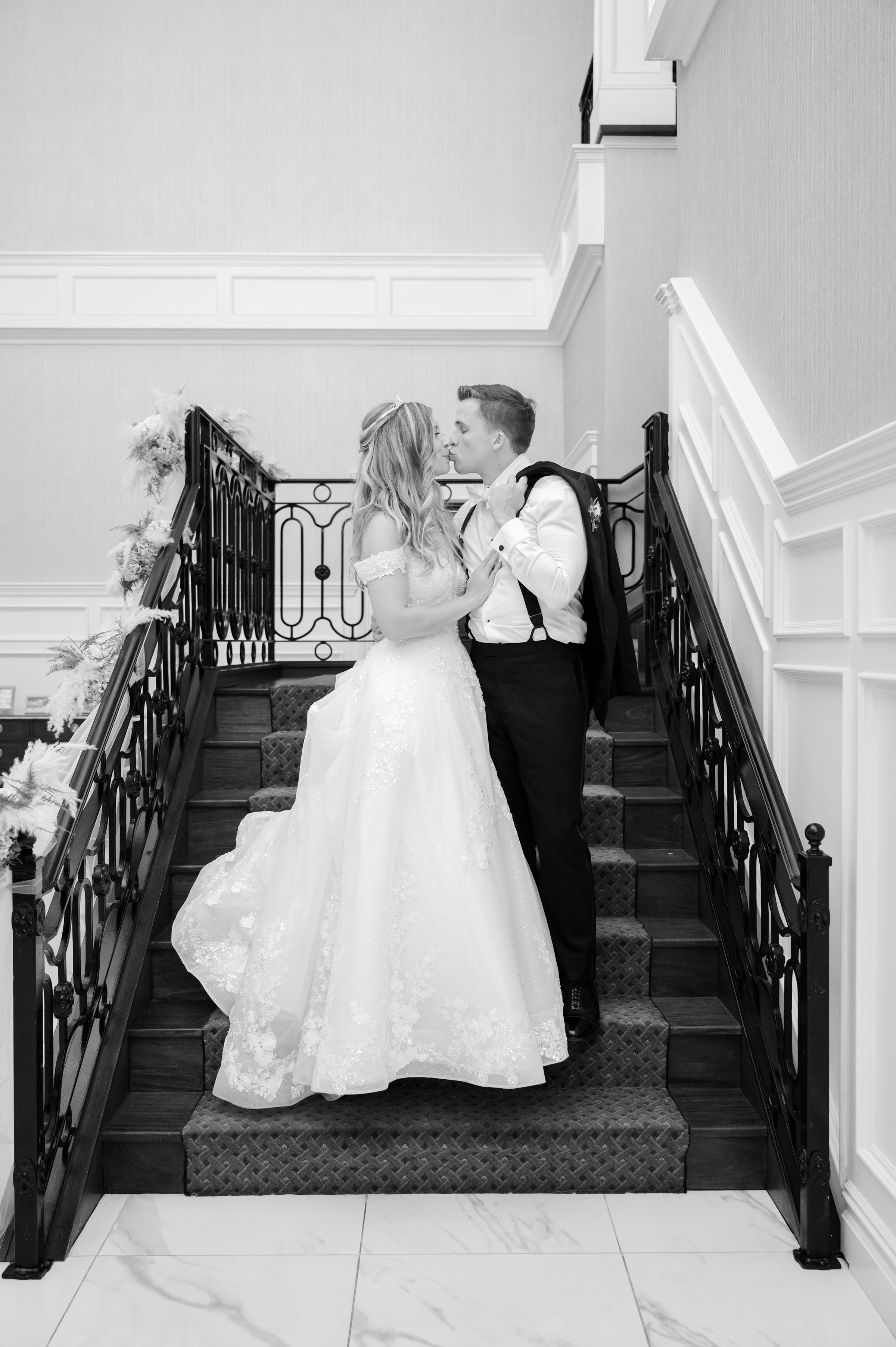 Bride and groom share a kiss on a grand staircase, surrounded by elegant white walls and black railings.