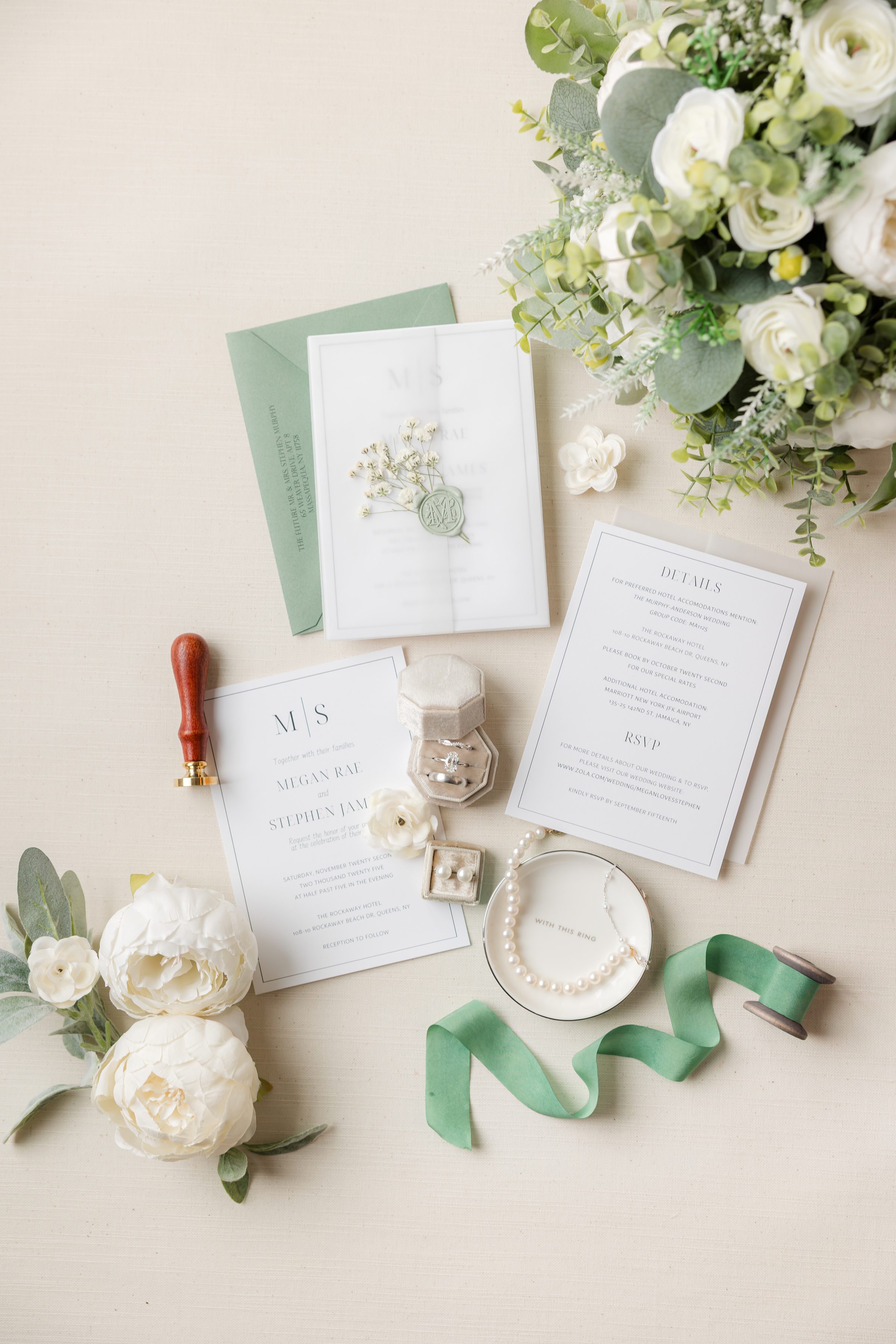 Wedding stationery flat lay featuring an invitation suite with envelopes, wax seal, rings, ribbon, and white floral arrangement on a neutral background.