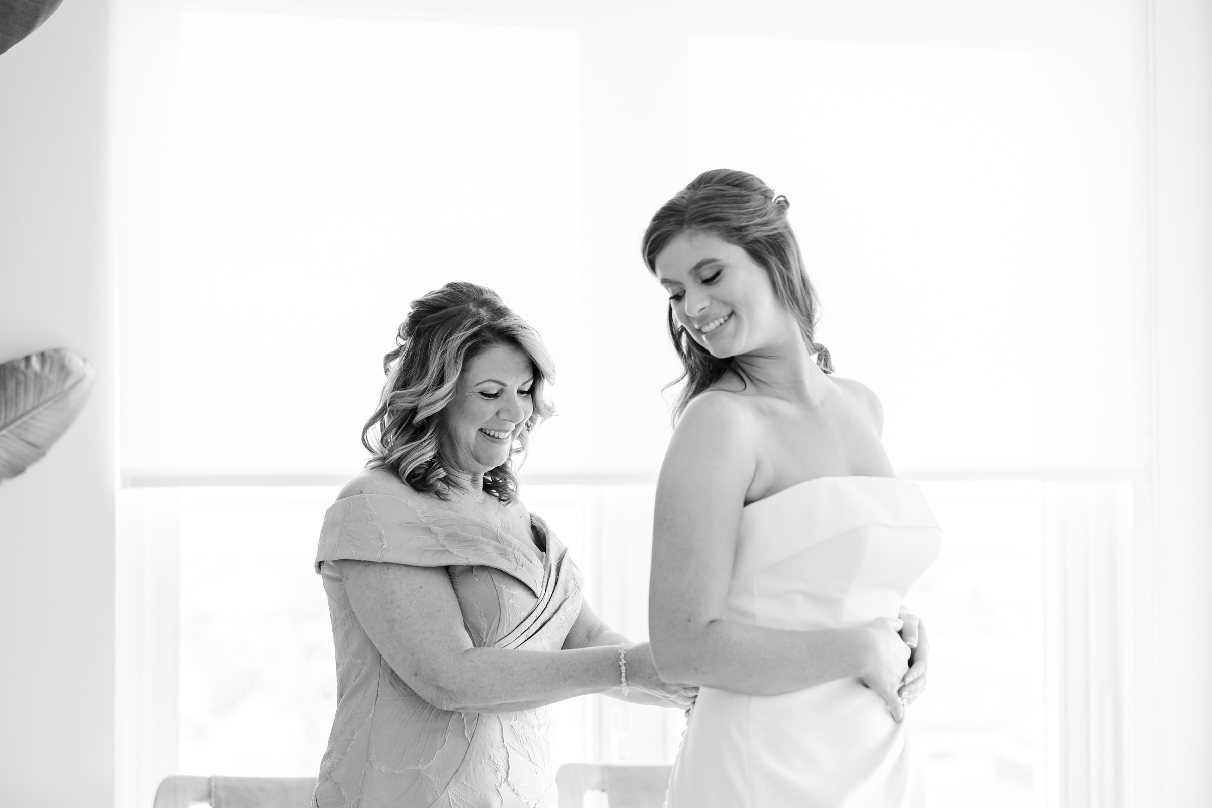 Bride smiling as her mother fastens the back of her wedding dress in a bright, softly lit room.