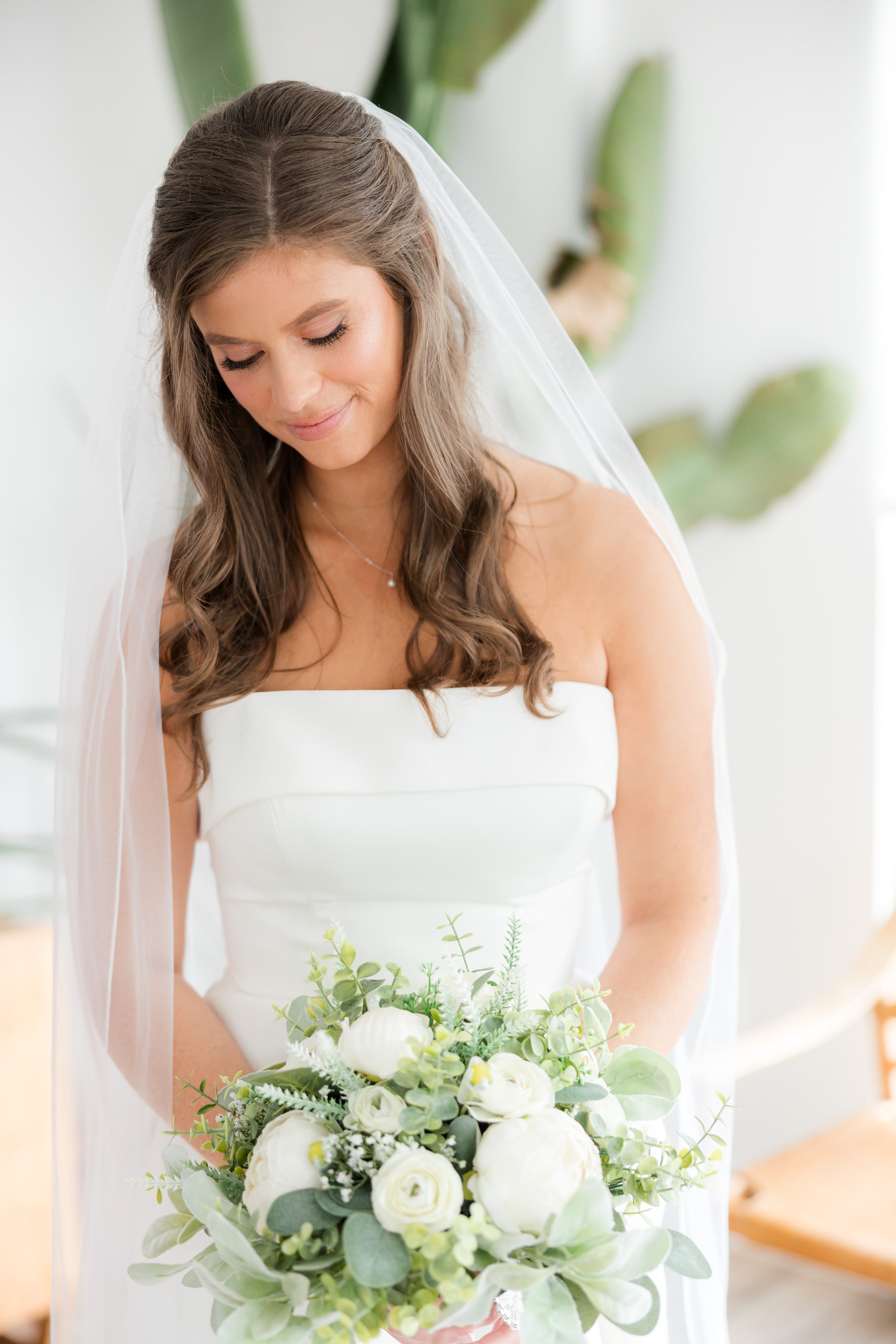 Bride holding a bouquet of white flowers and greenery, looking down softly.