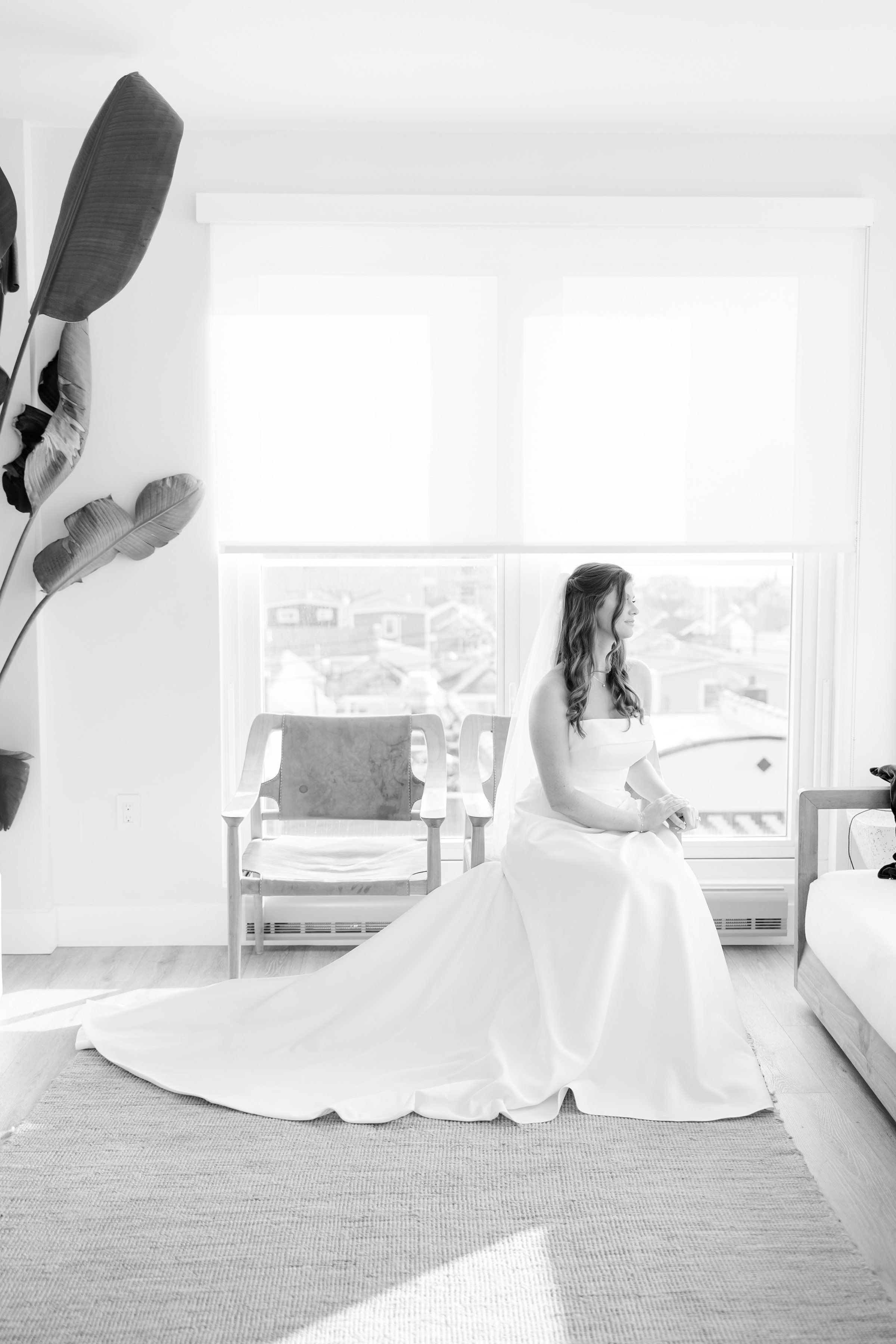 Bride sitting in a bright room by a large window, wearing a strapless gown and veil.