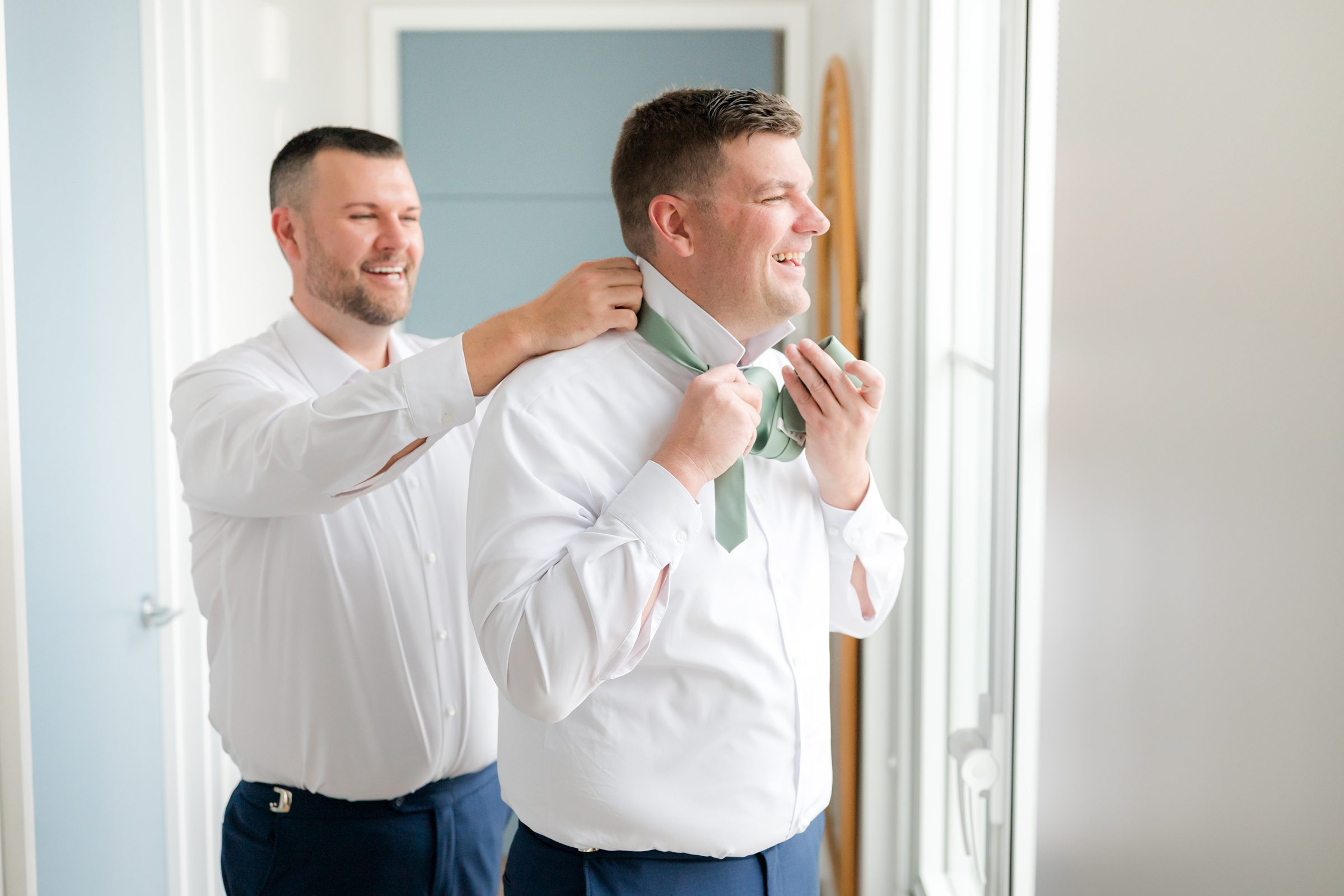 Groom smiling as a groomsman helps tie his light green necktie by a window.