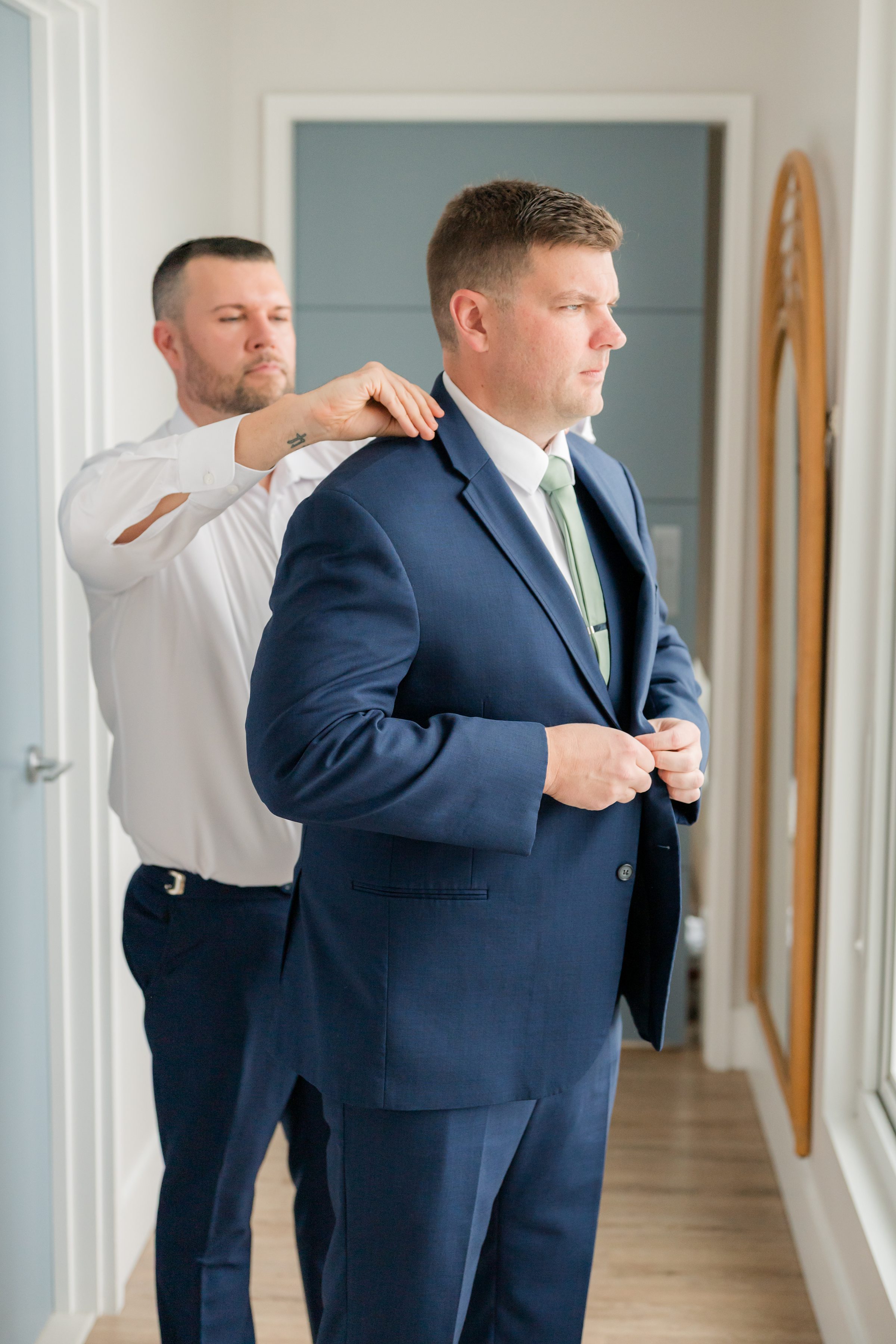 Groom getting ready in a blue suit as a groomsman adjusts his jacket in a bright room.