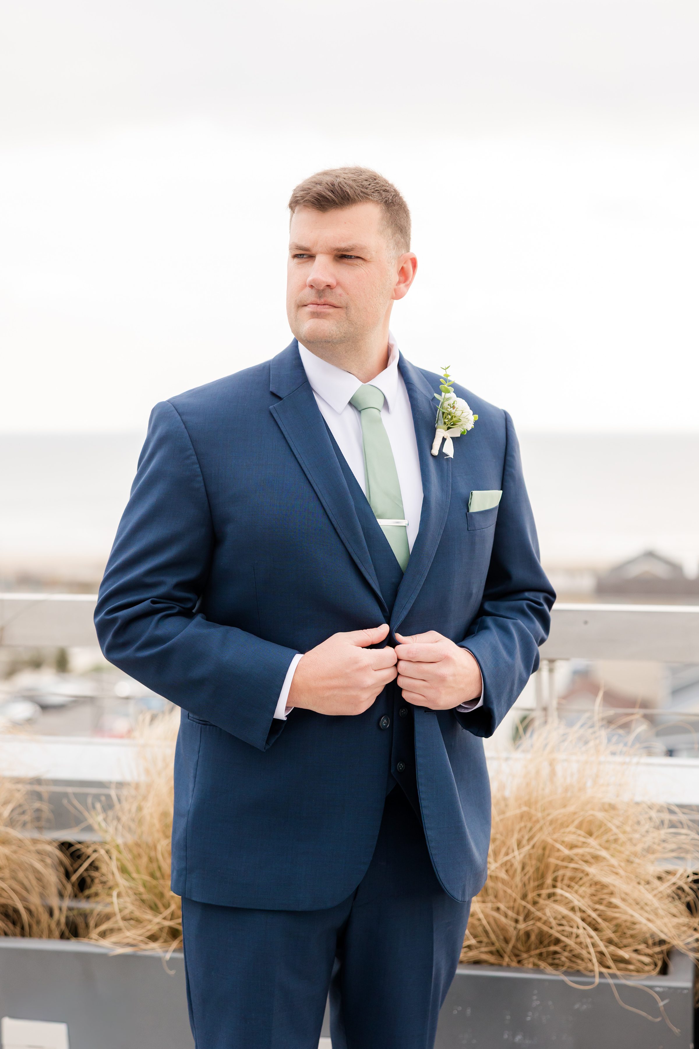 Groom in a blue suit with a light green tie standing outdoors, looking off to the side.